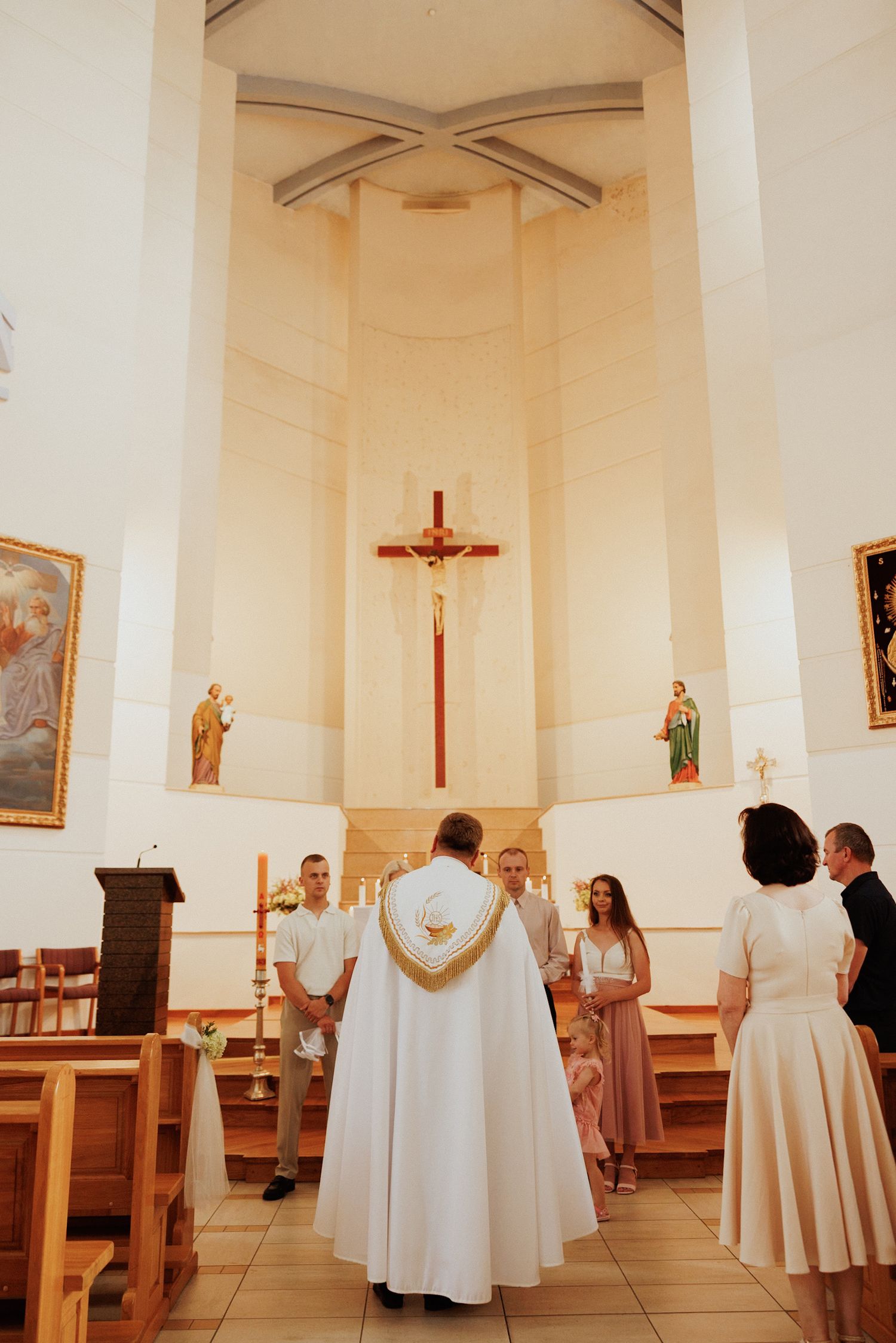 White-robed figures gather inside a church sanctuary with wooden pews and religious artwork on cream-colored walls.