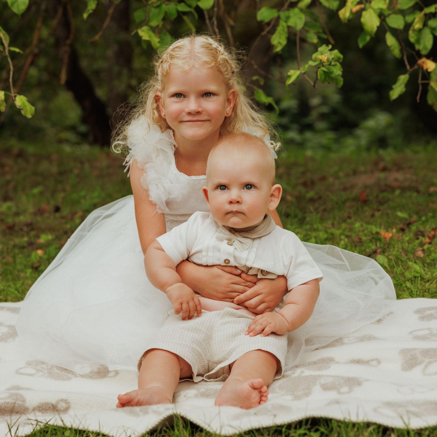 Children sitting together on white blanket in lush garden setting.