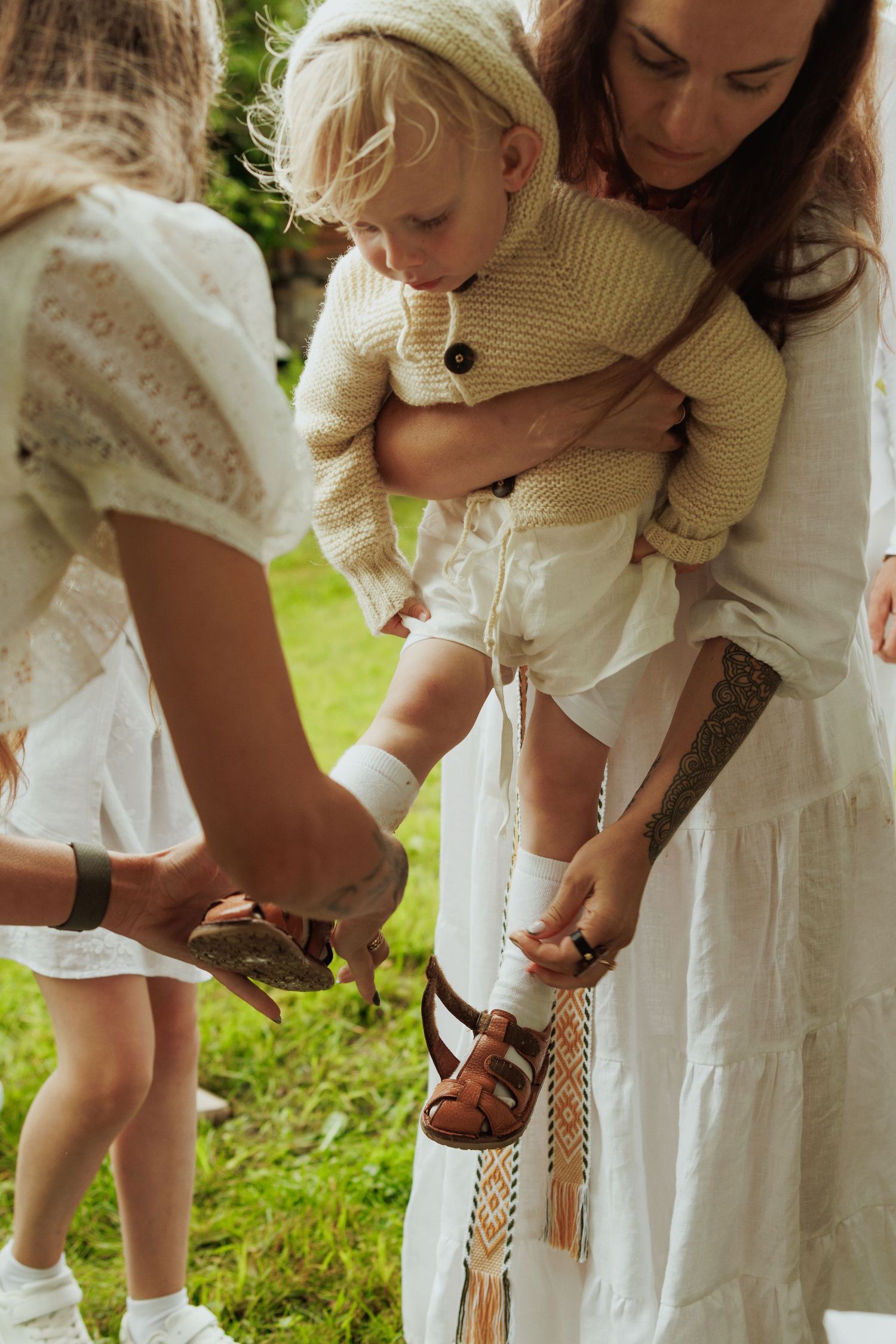 Mother in white dress holding toddler while woman helps put on sandals in a garden setting.