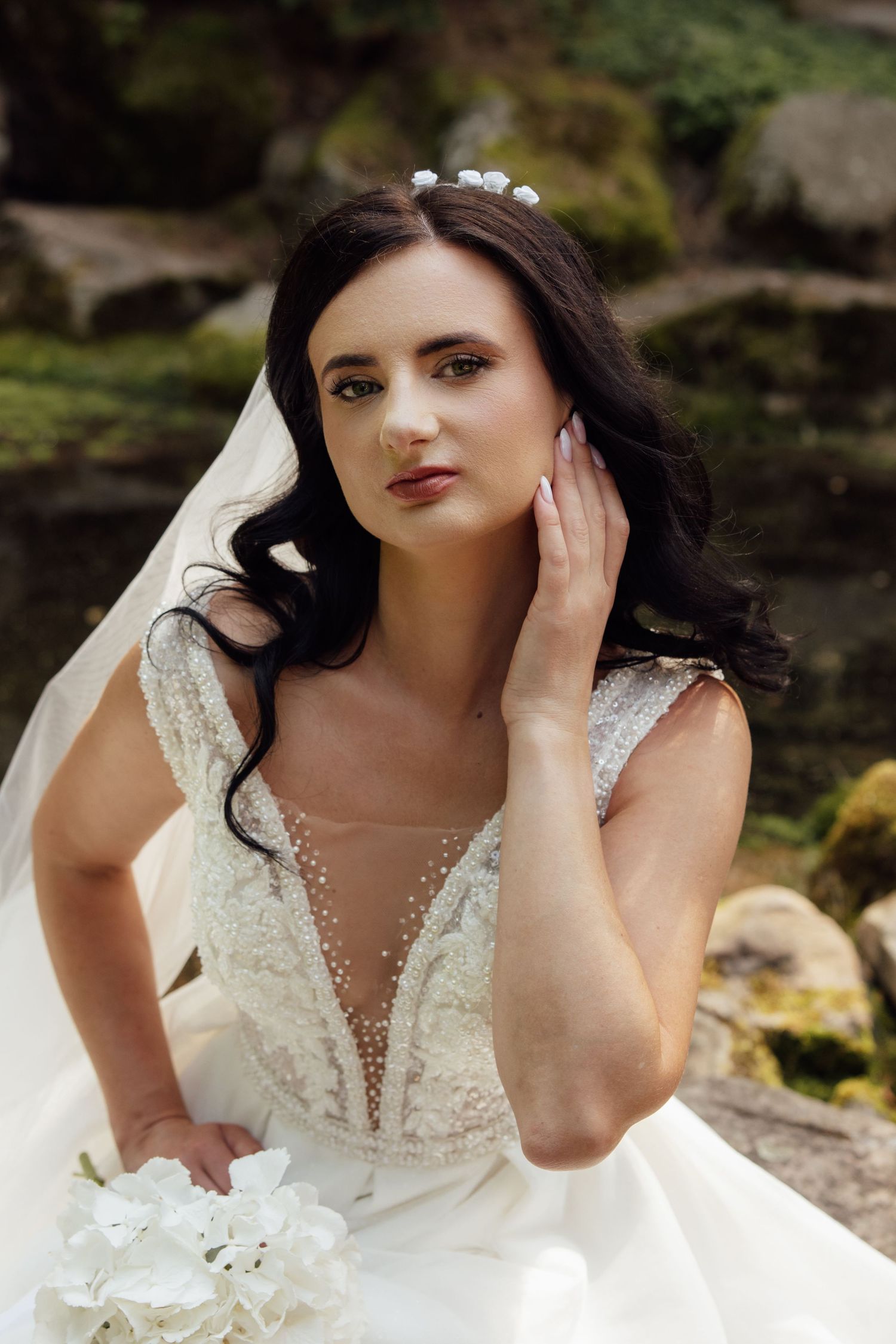Beautiful dark-haired bride in beaded lace wedding dress with veil and floral hair accessory poses outdoors.