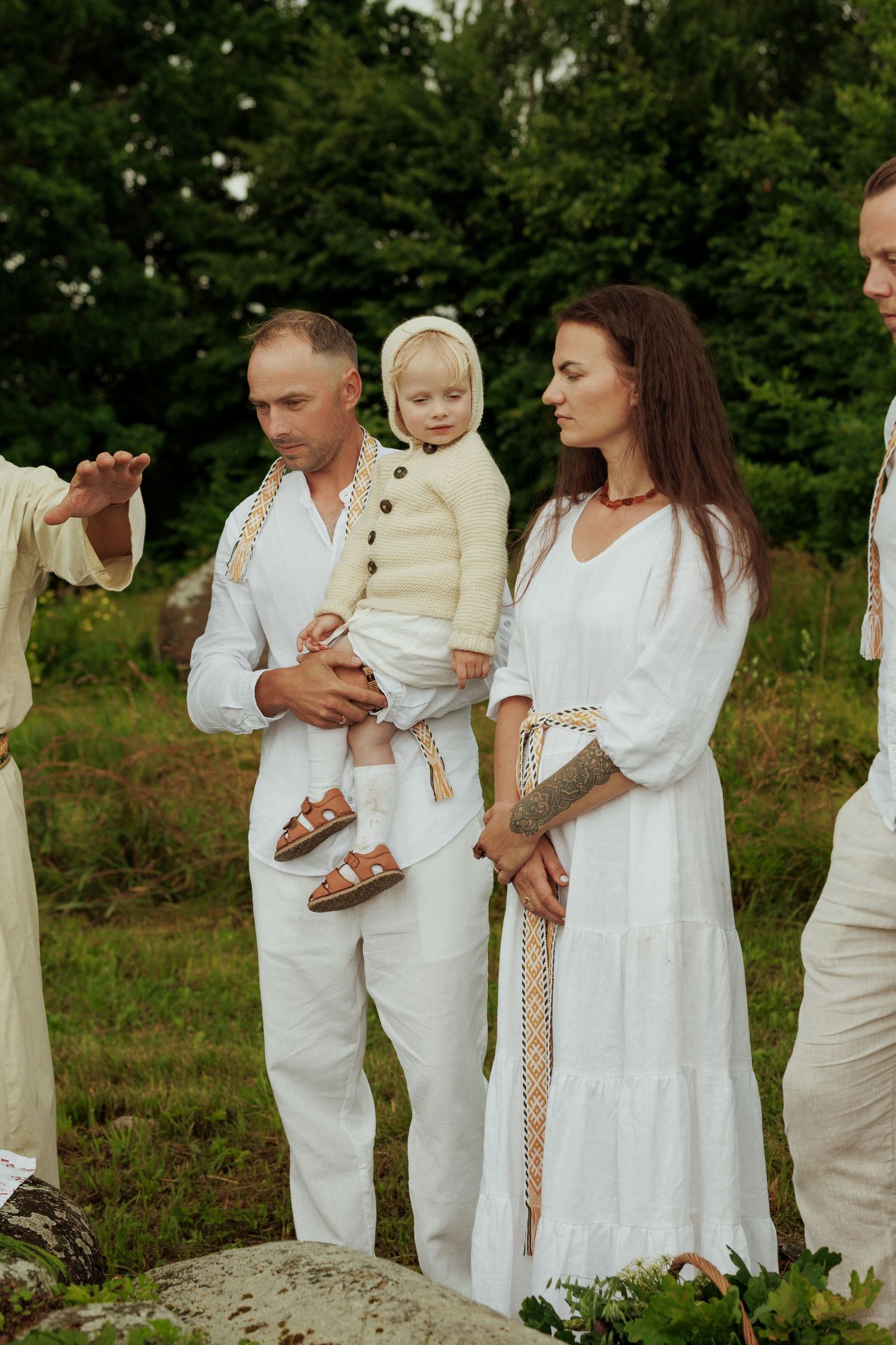 Family dressed in white traditional clothing holding a baby at an outdoor nature ceremony.