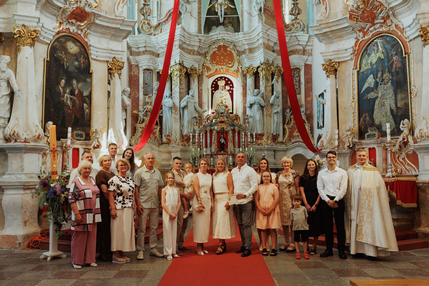 Wedding party gathered inside an ornate baroque church with red drapes and gilded decorative elements.