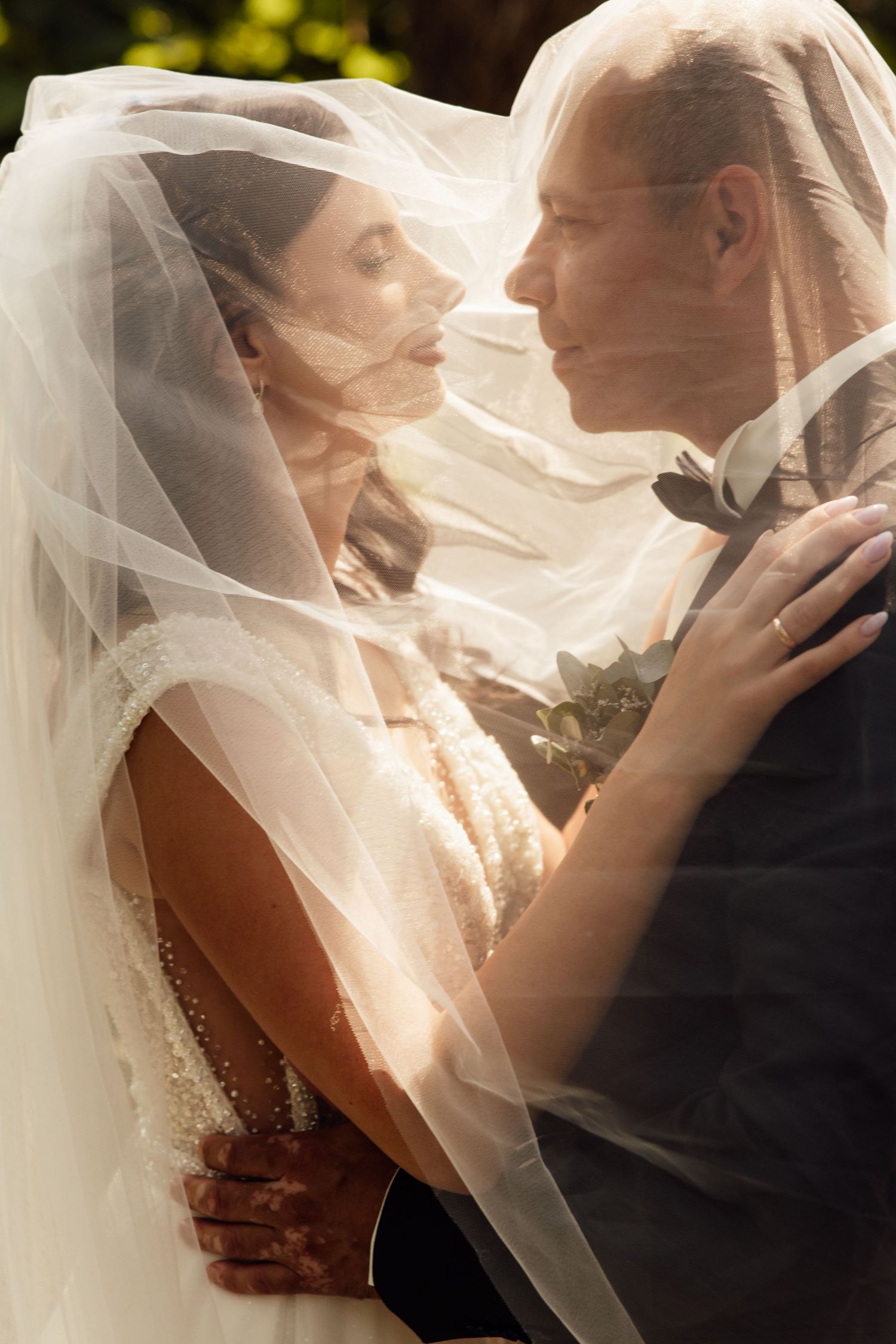 Bride and groom share an intimate moment under a flowing white veil, dressed in wedding attire outdoors.
