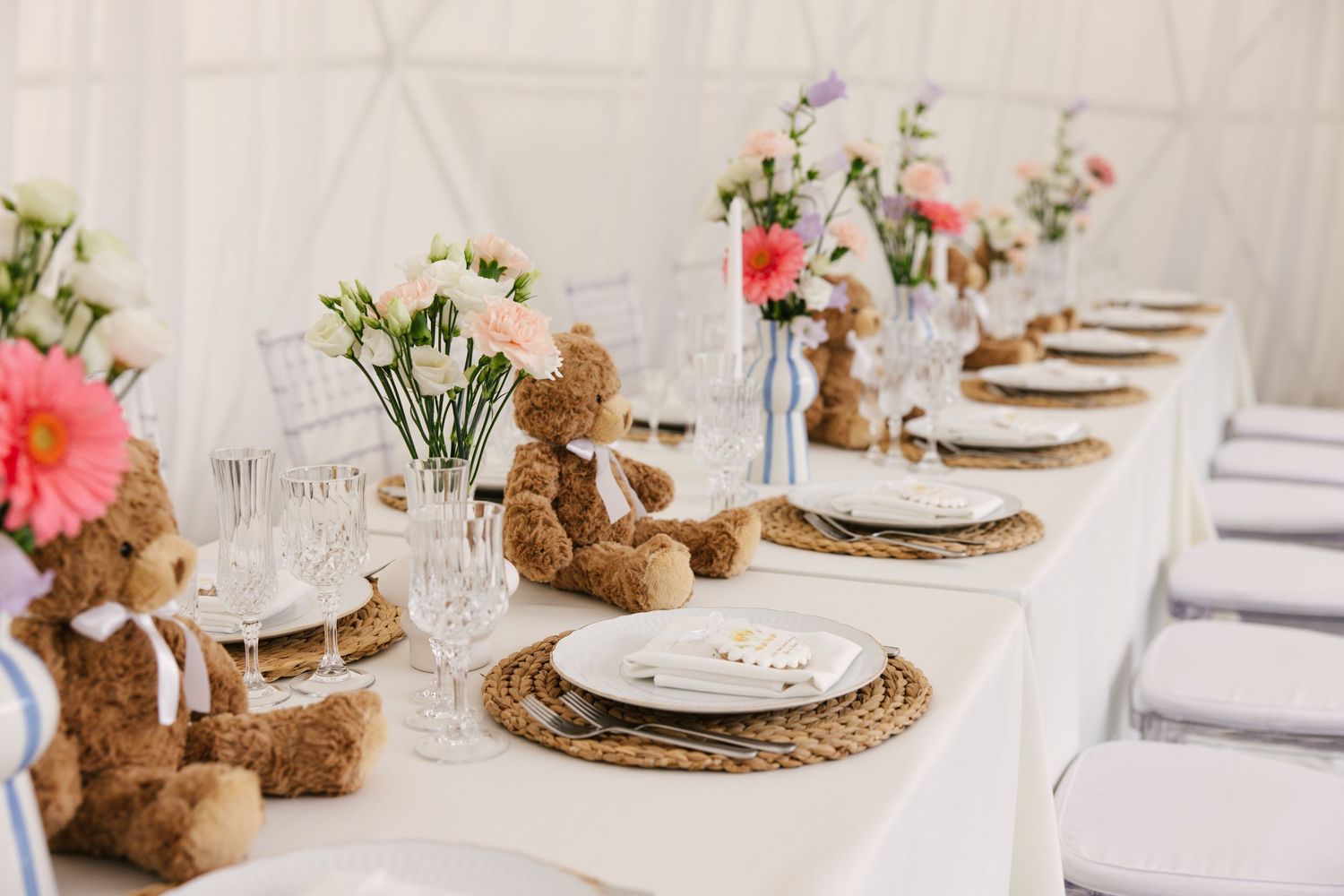 Long dining table decorated with teddy bears and pink flowers on woven placemats.