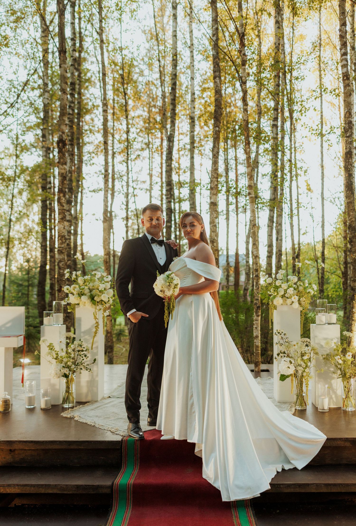 Wedding ceremony setup in a birch forest with white pedestals and floral arrangements along a red carpet.