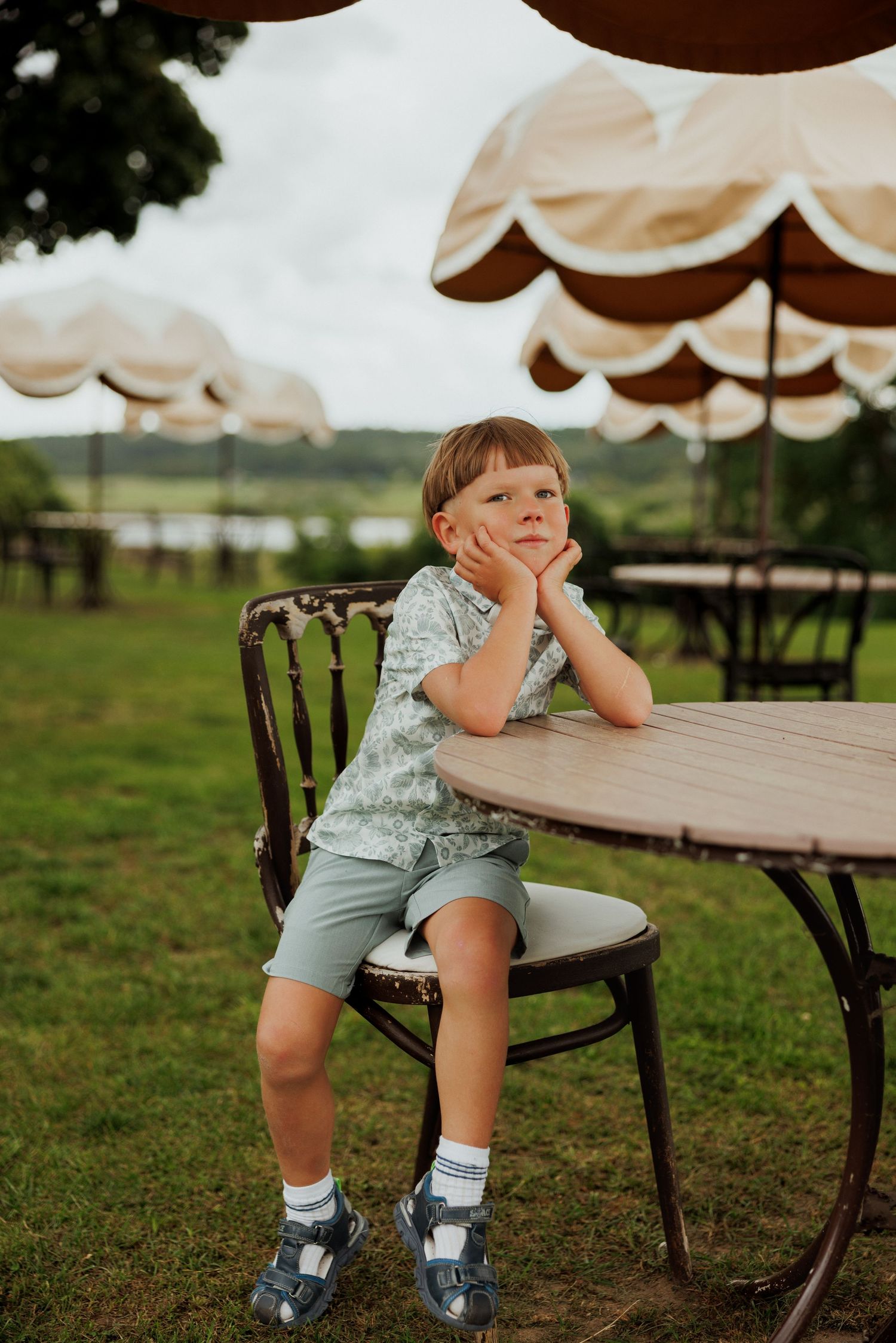 Child in casual summer clothes sitting at an outdoor table under beige umbrellas in a garden setting.