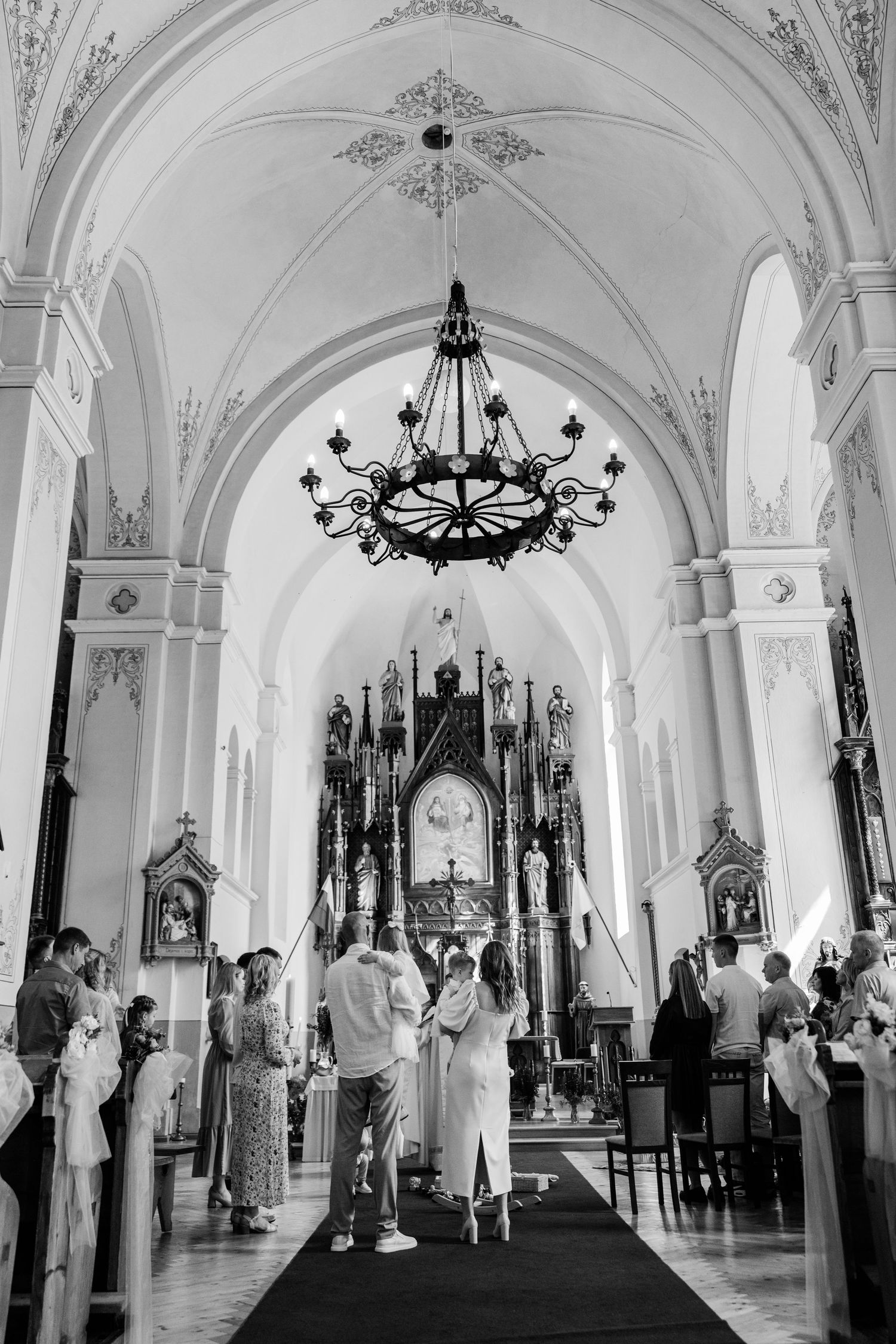 A panoramic view of a white church interior with arched ceilings, chandeliers, and people gathered for a ceremony.