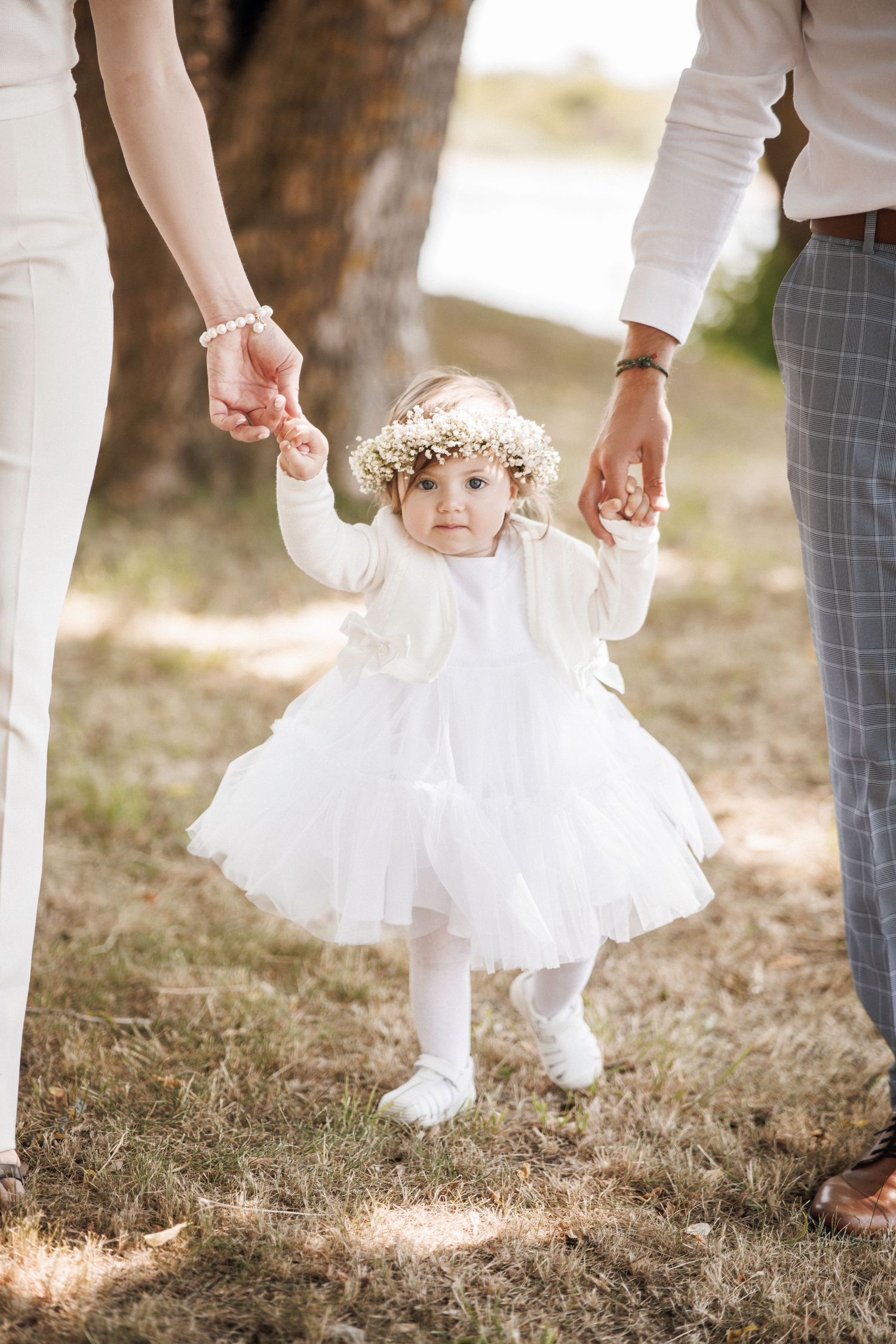 A child in a white dress and flower crown holds hands with adults wearing white and gray outfits in an outdoor setting.