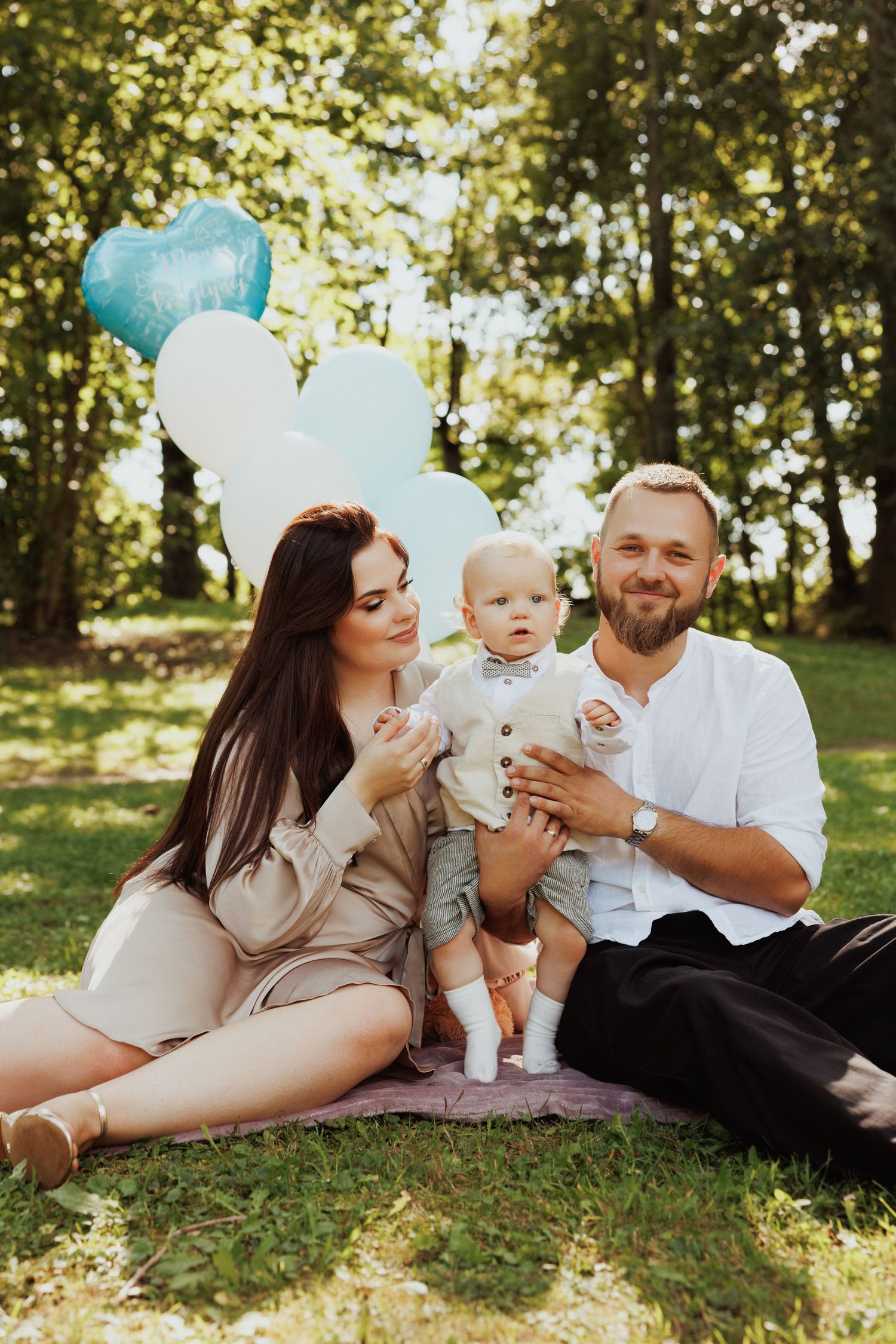 Family laying on grass in a sun-dappled park setting with blue balloons during a casual outdoor photoshoot.