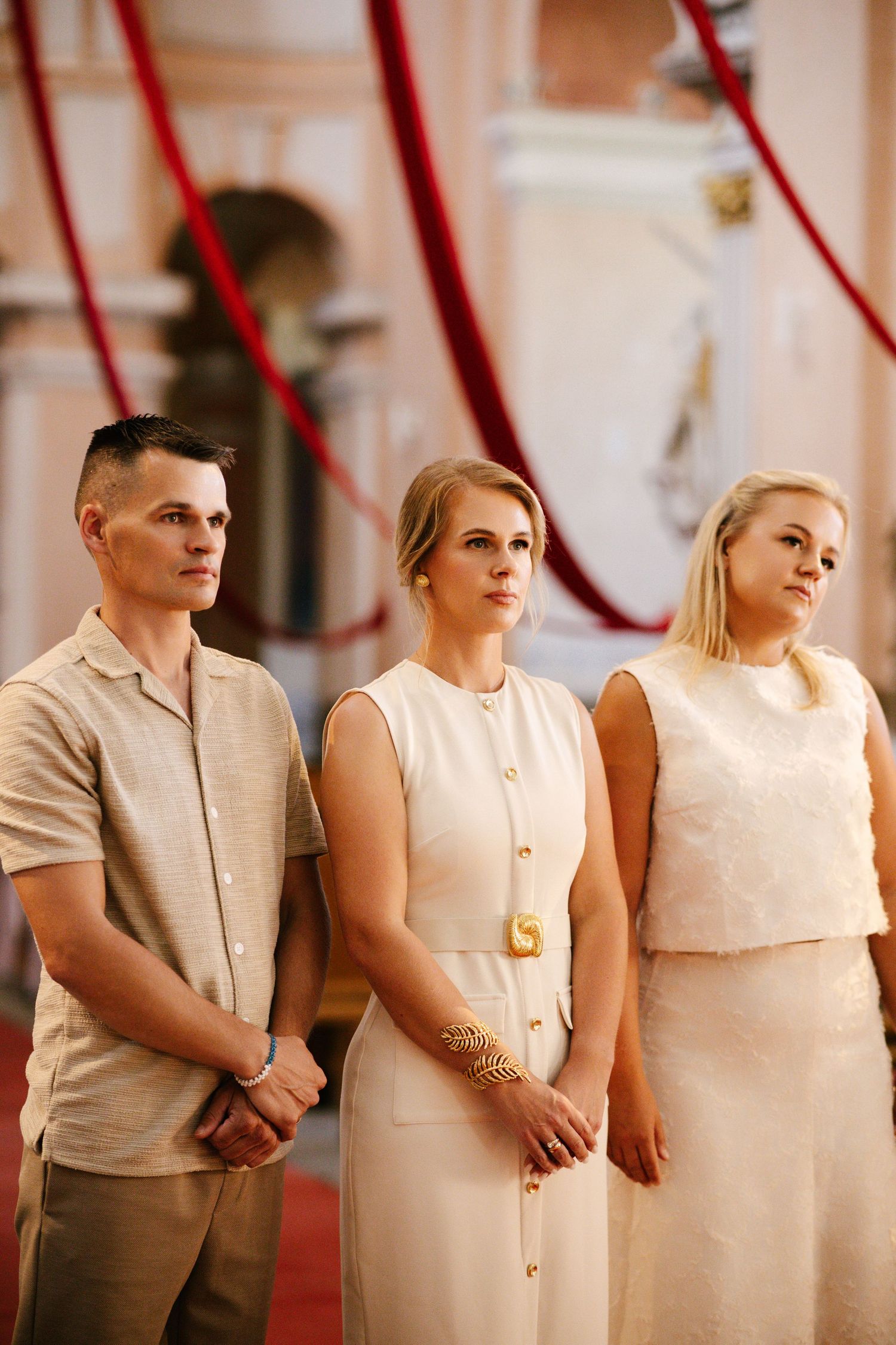 Formal event photograph with three people in neutral-toned attire standing against a pink wall.
