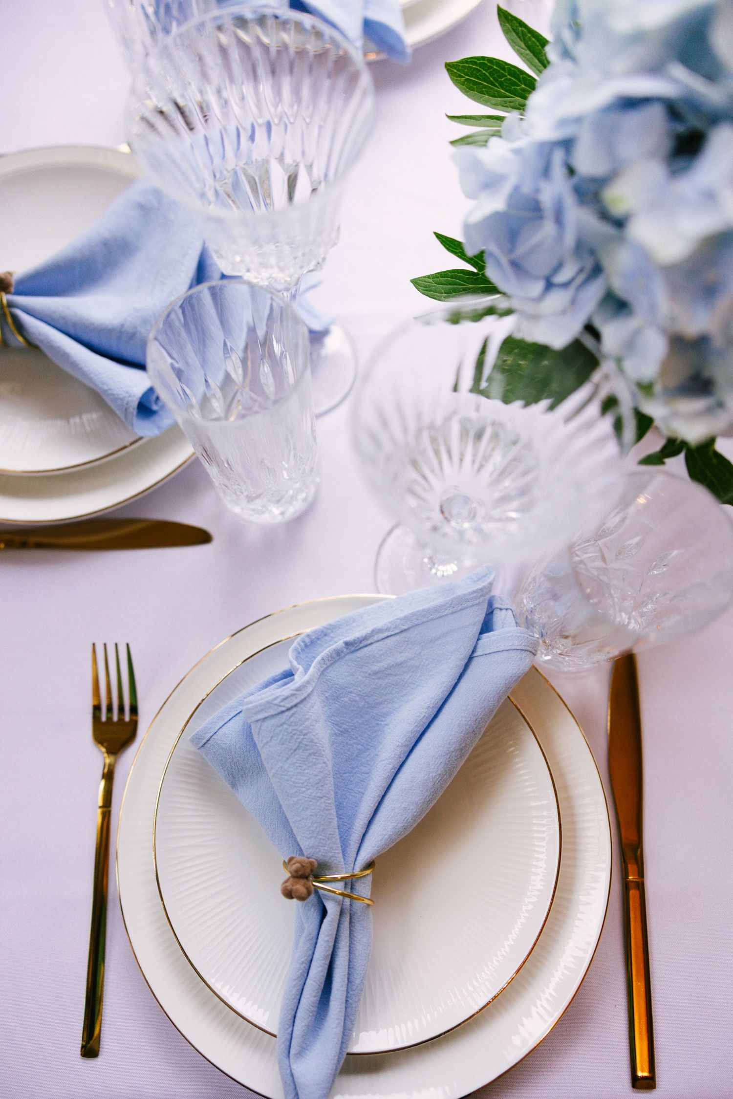 Formal place settings with light blue napkins, gold cutlery, and crystal glasses on white tablecloth.