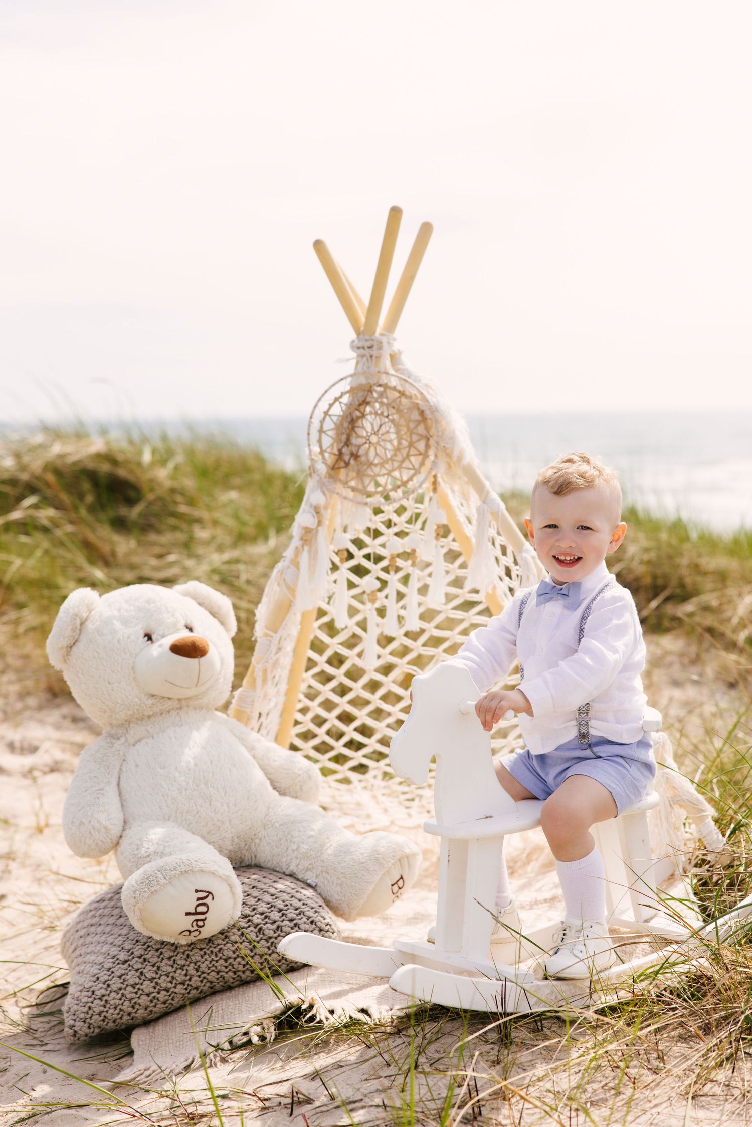 Beach setup with white teddy bears and decorative teepee on sandy dunes.