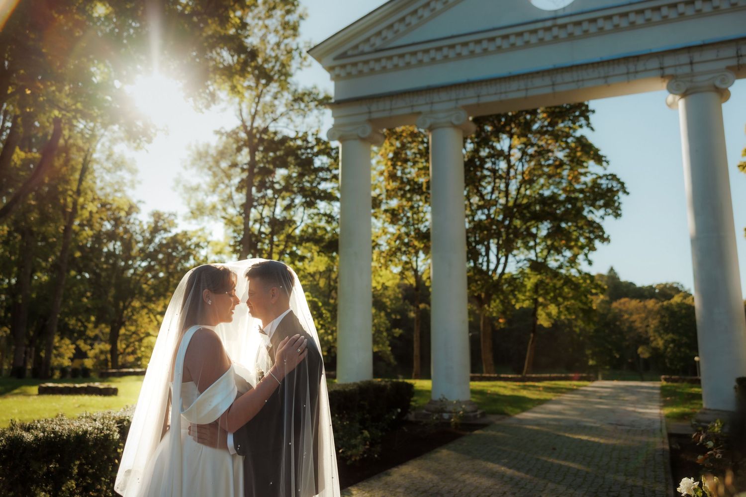 Wedding ceremony setting with white columns and golden sunlight filtering through trees.