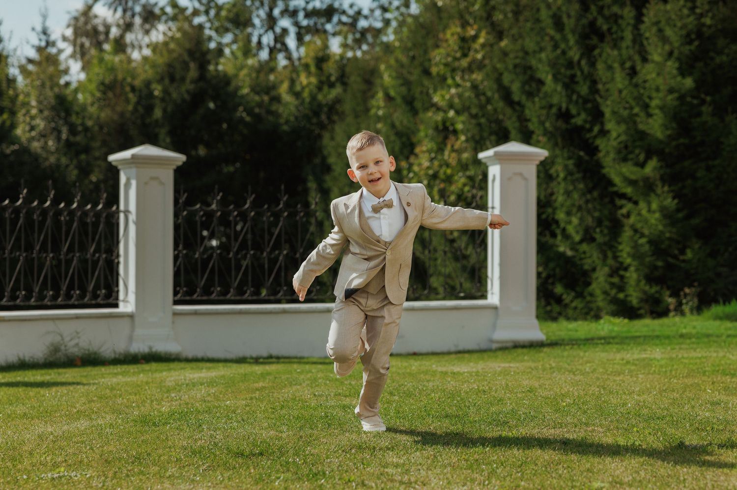 A child plays outdoors in a sunny garden setting with white fence posts and green trees in the background.