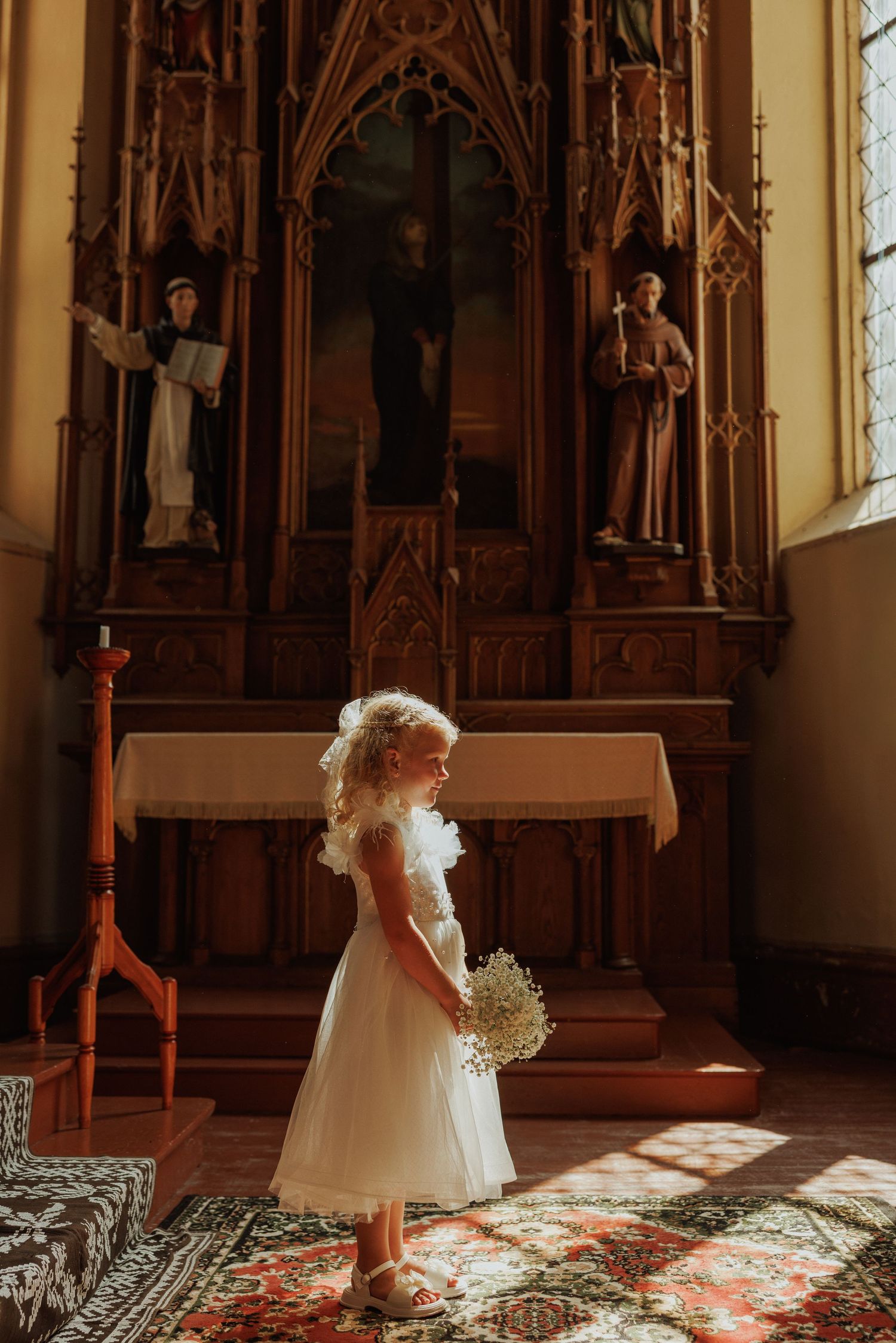 Sunlight streams through church windows onto an ornate carpet as someone in a white dress stands in the glow.