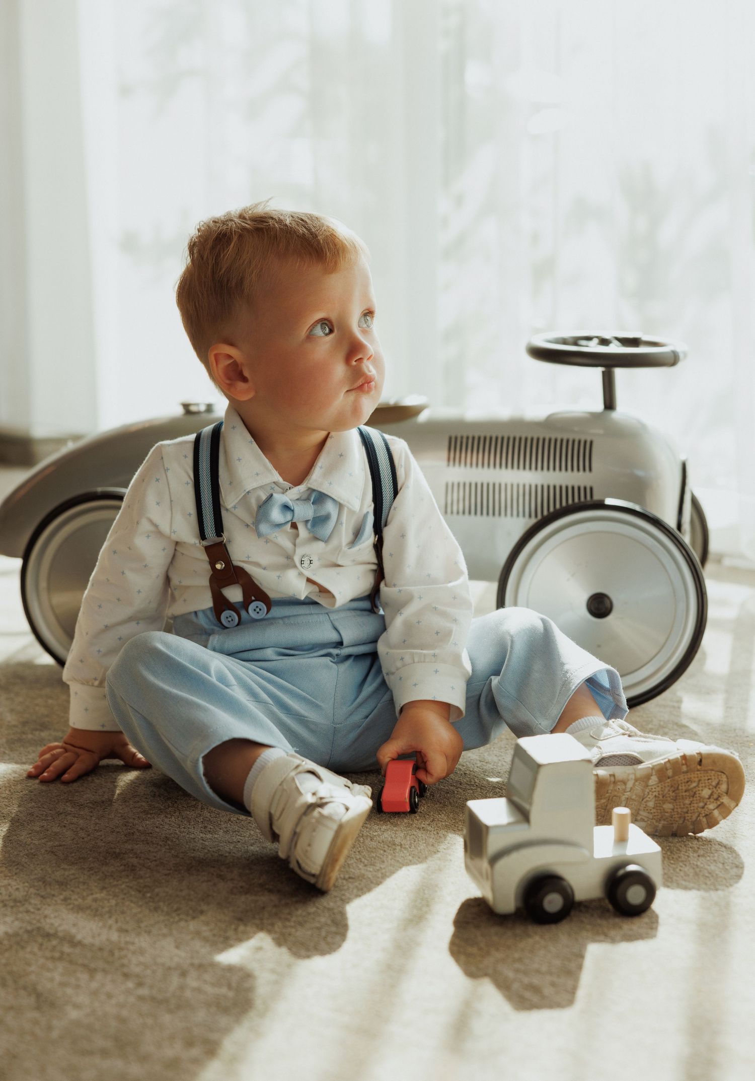 A baby in blue overalls sits on a carpet playing with toy cars while wearing a light colored dress shirt.