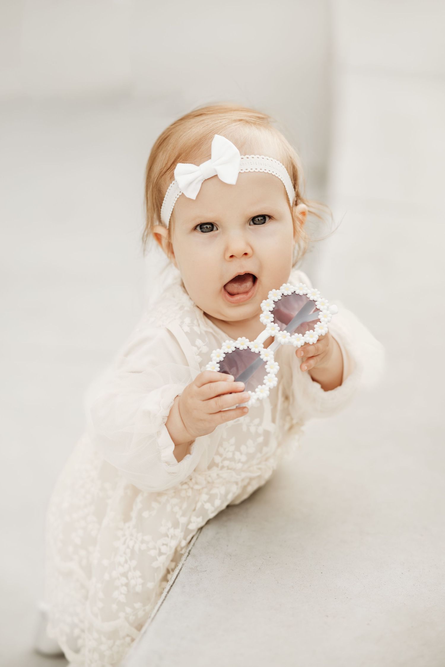 Baby in white lace dress holding a flower while lying on light background.