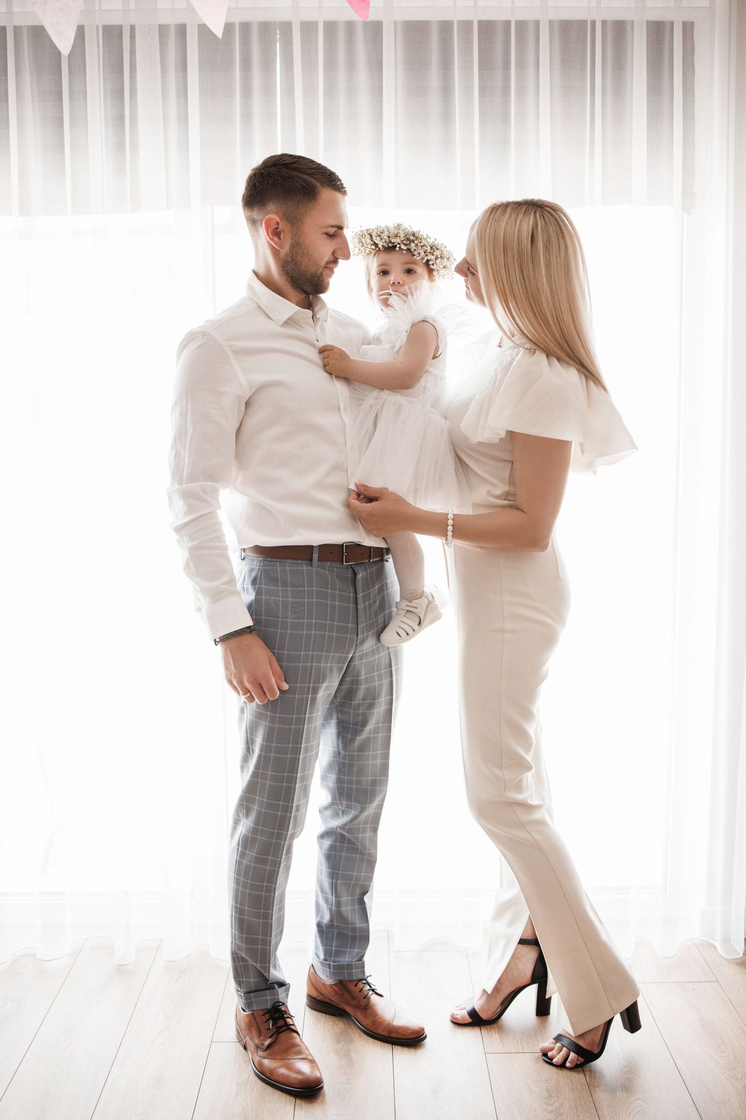 A couple in white outfits dances together in front of sheer curtains in a bright room.