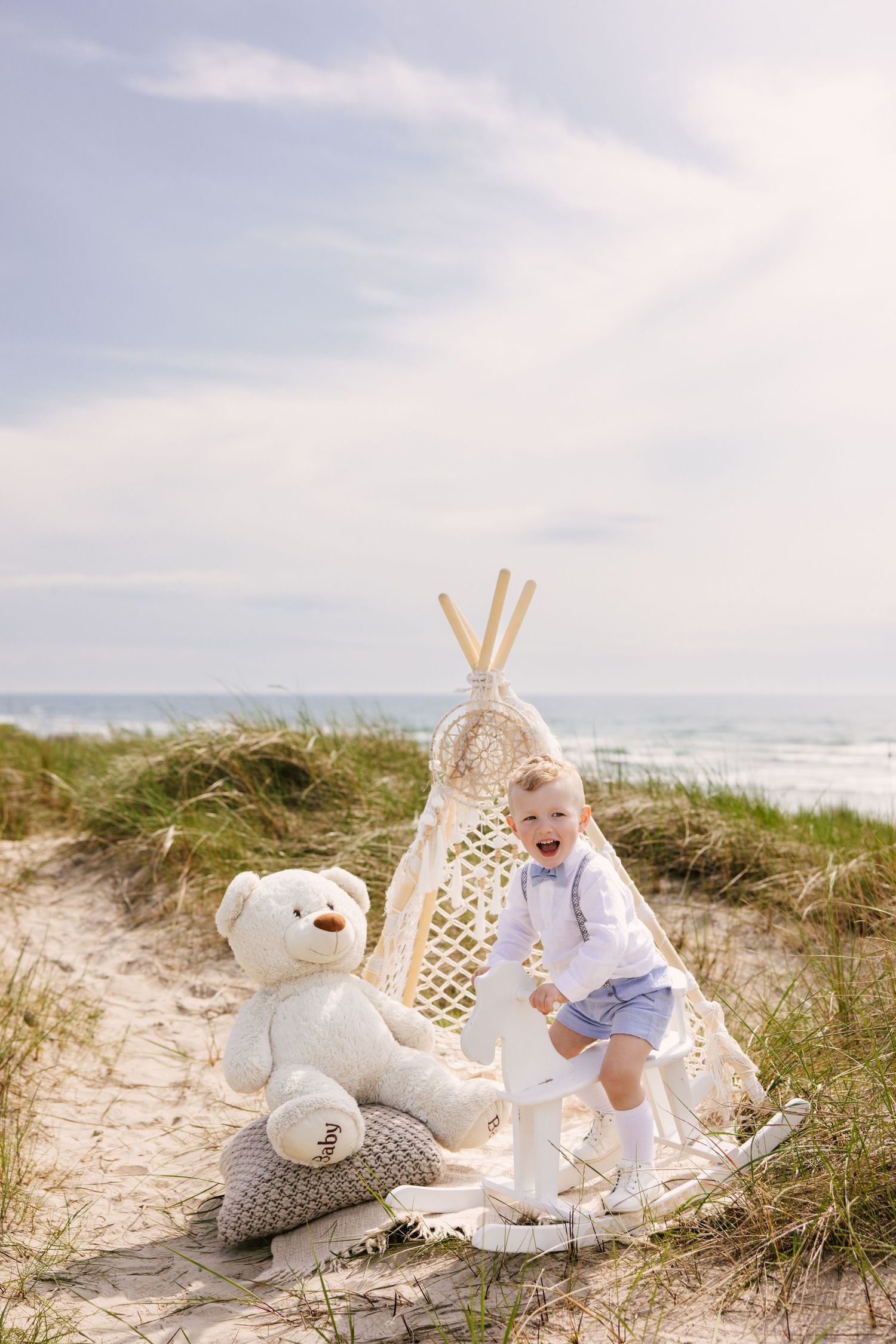 A small child plays next to a decorative teepee and teddy bear on a sandy beach with ocean waves in the background.