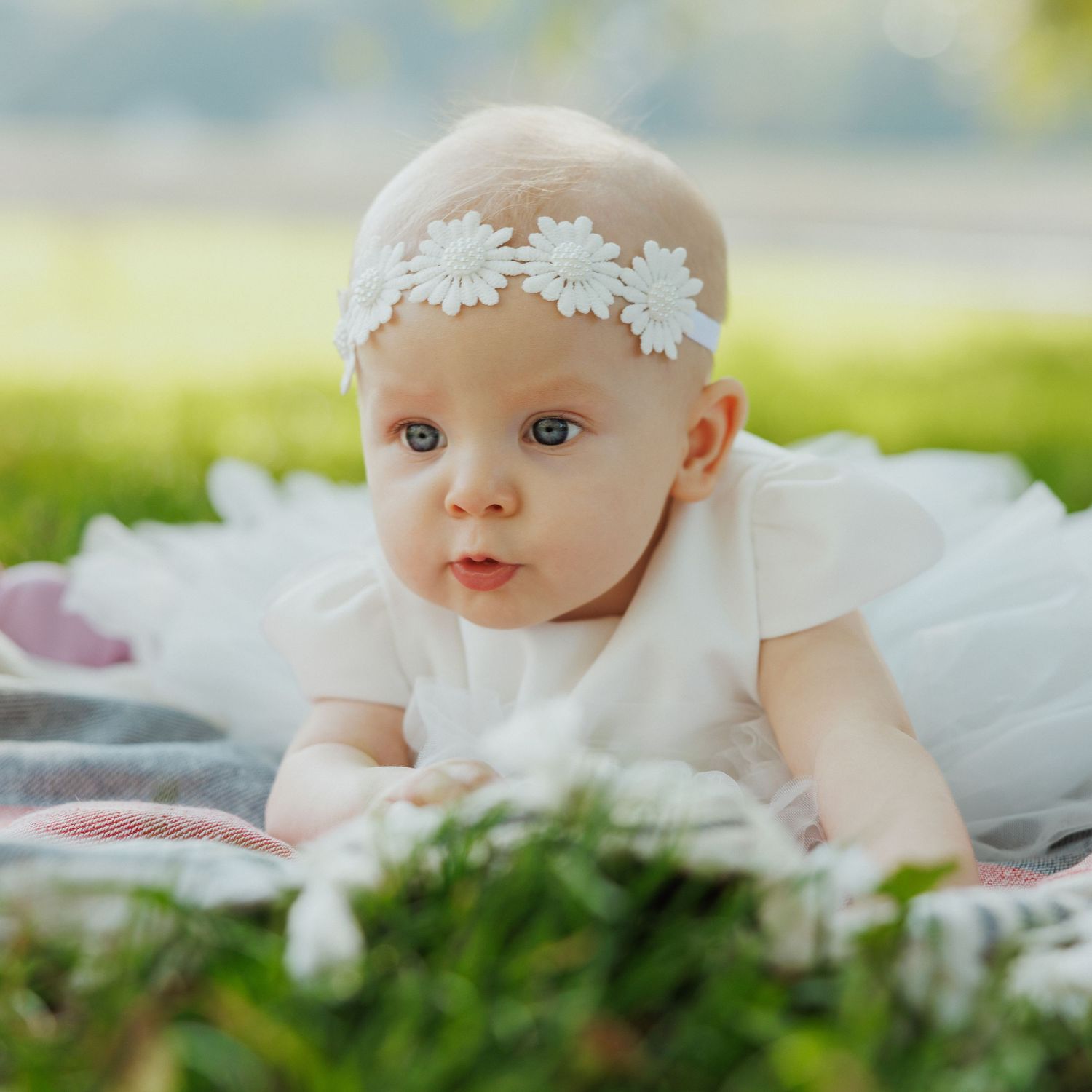 Baby wearing white lace headband lying on grass in outdoor portrait session.