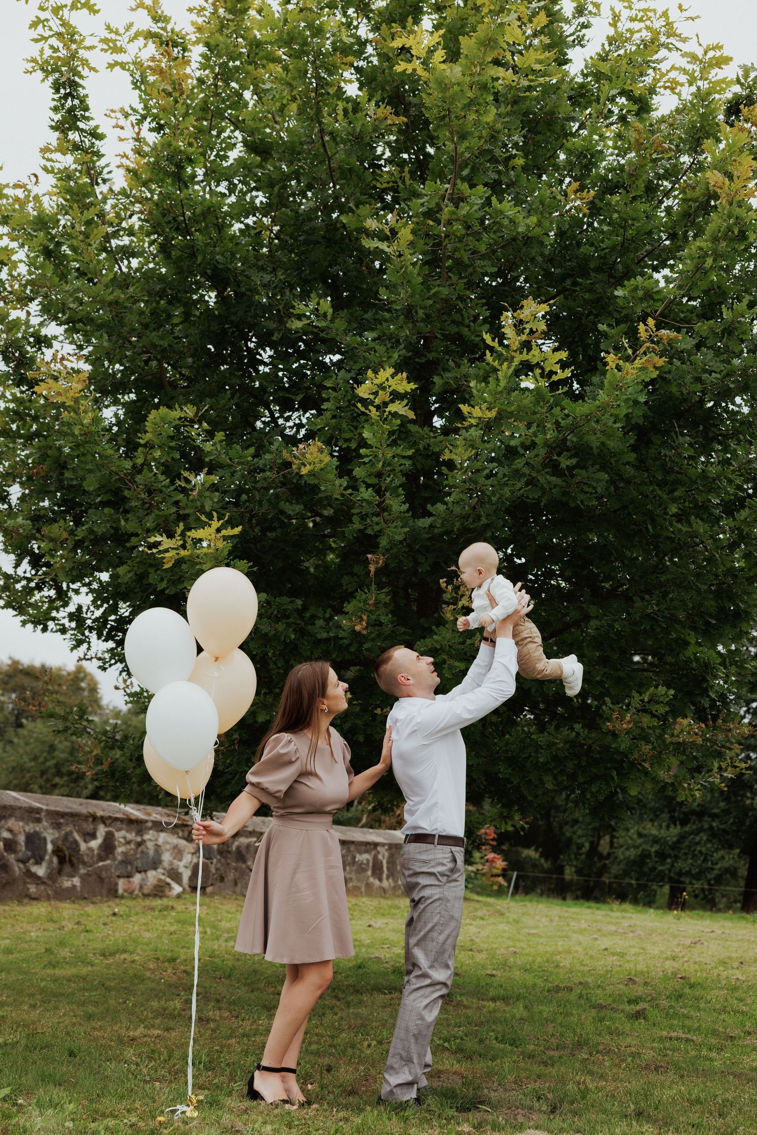 A couple releases white balloons into the air in a grassy field surrounded by trees during a photo shoot.