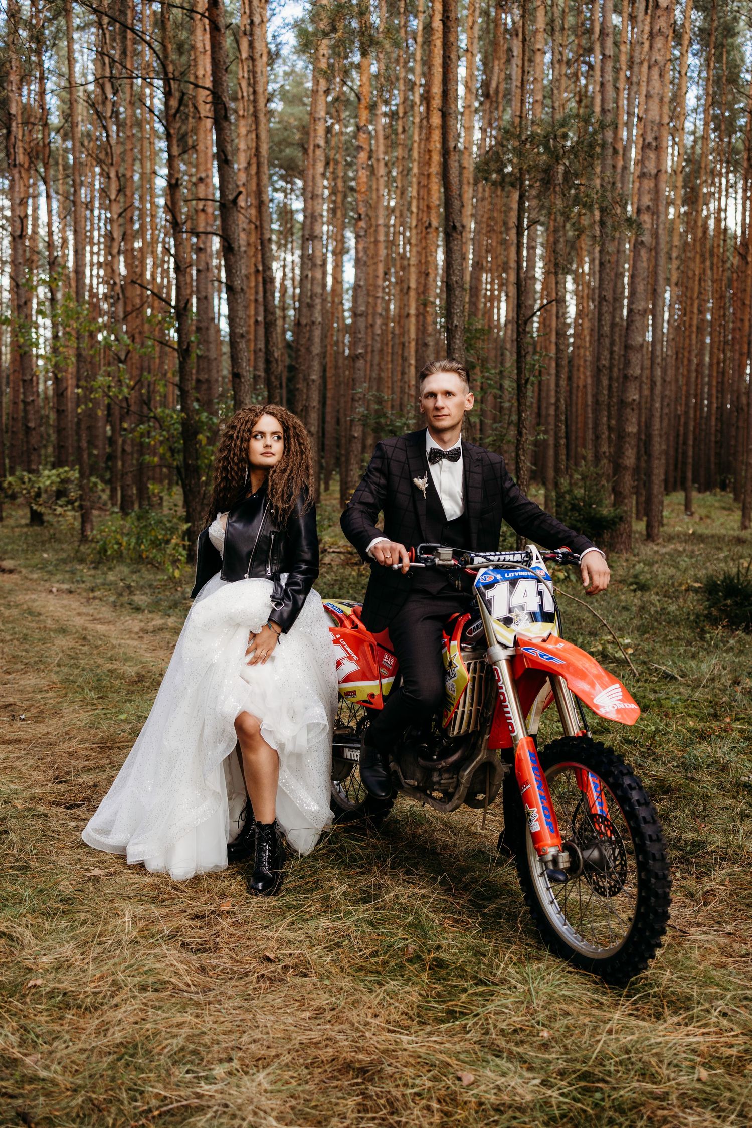 Wedding couple poses with a red dirt bike in a serene pine forest setting.