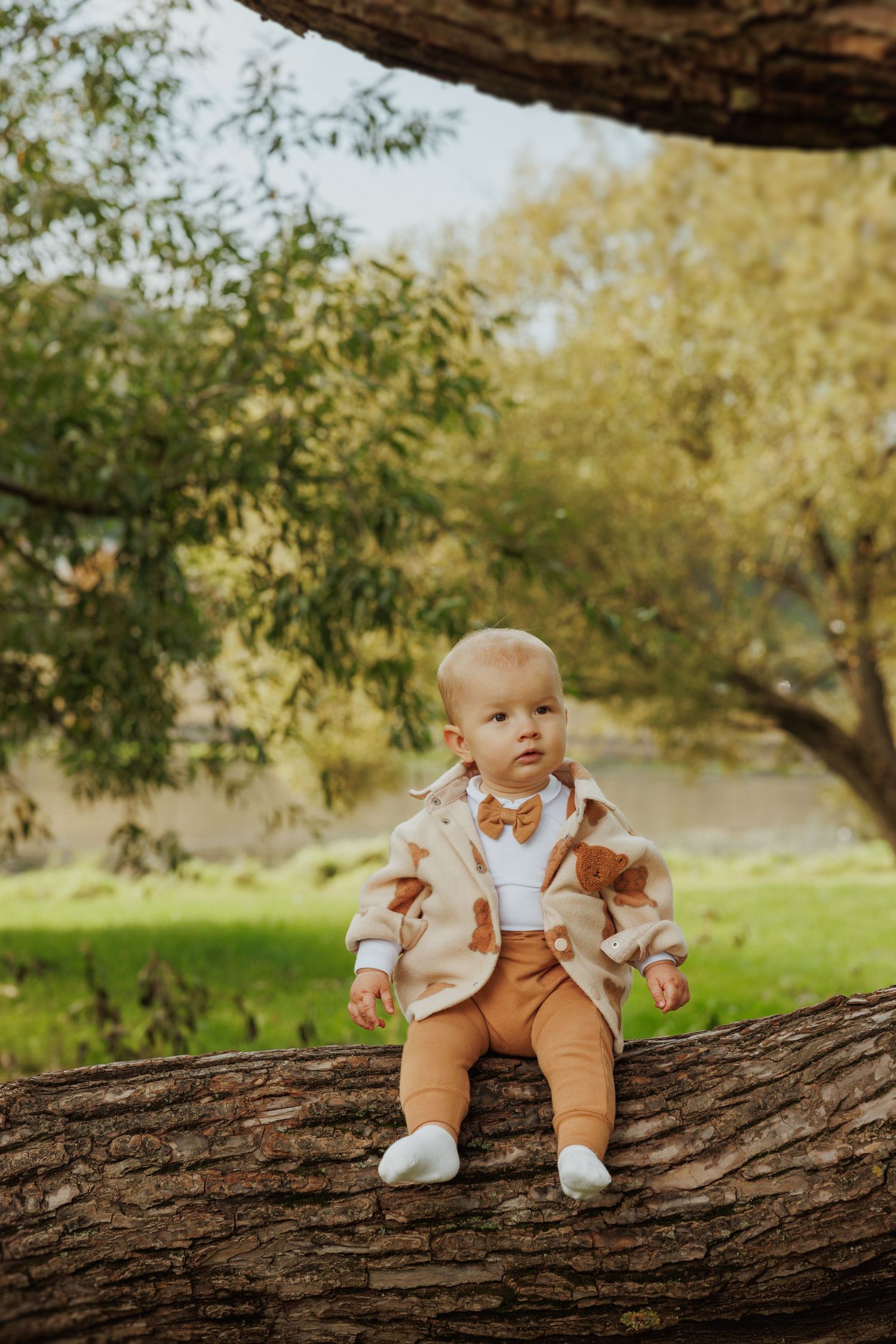 A baby wearing a floral jacket and brown pants sits on a tree branch in an autumn garden.