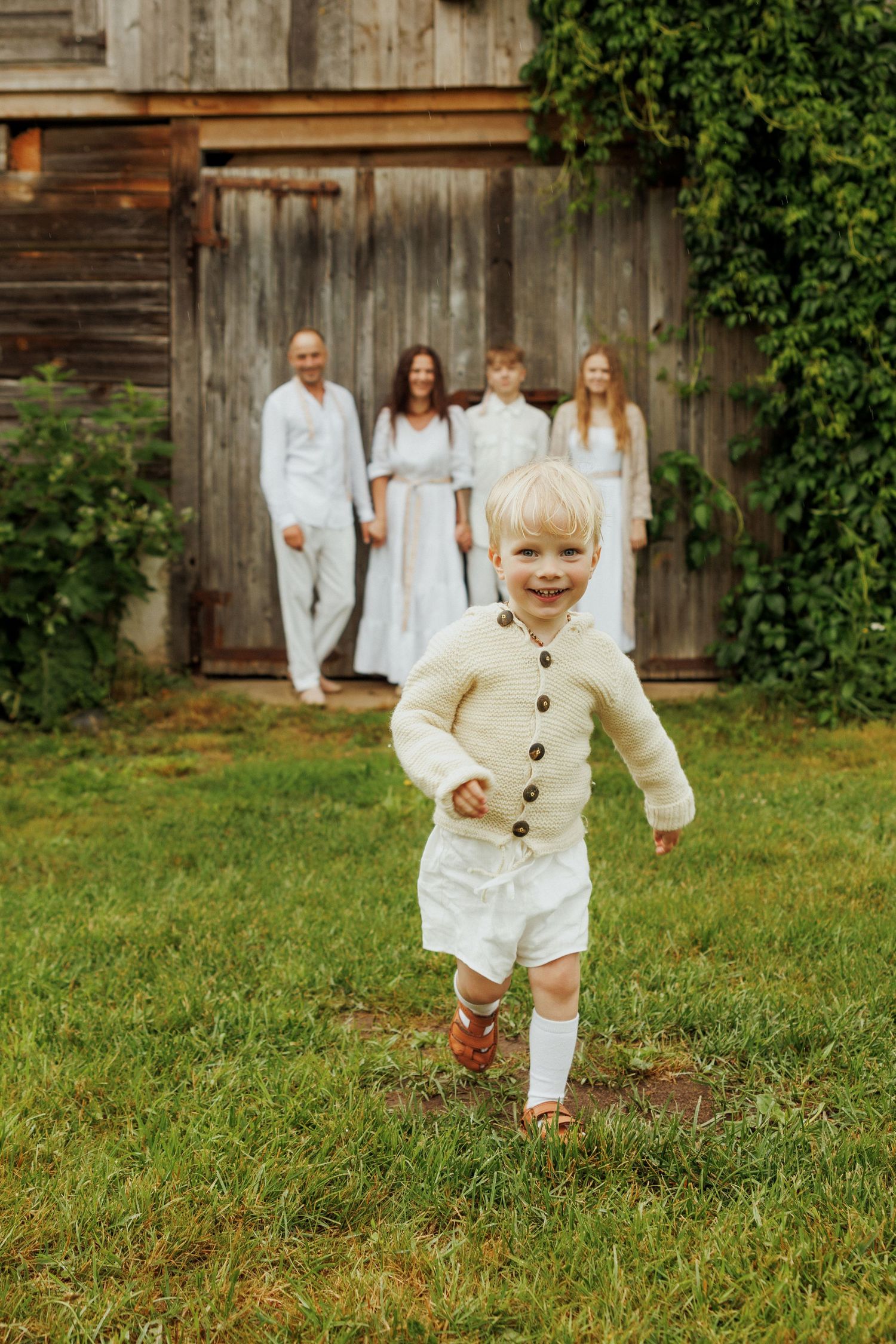 Smiling toddler in cream cardigan running on grass with family posed near rustic wooden barn door.