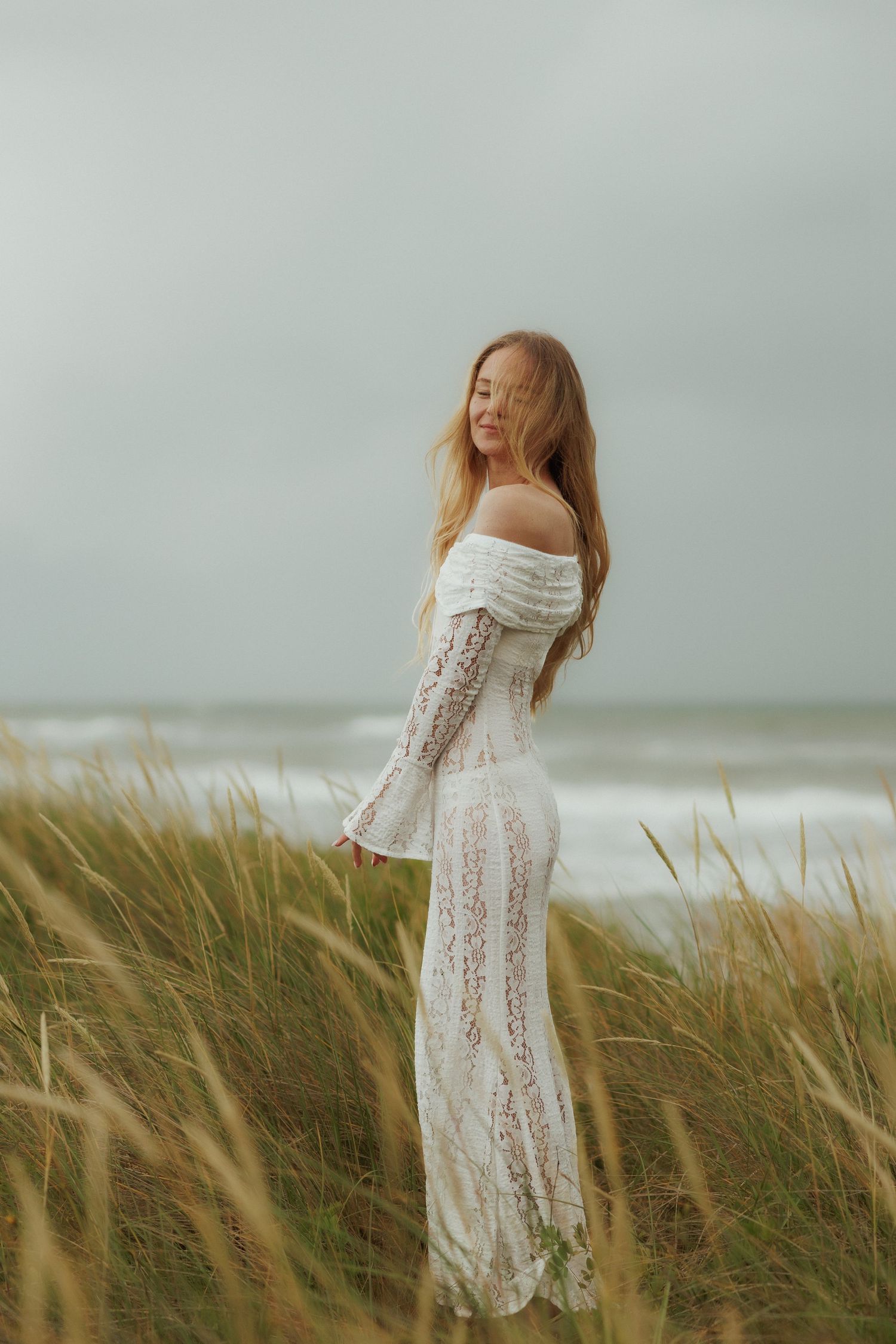 Elegant woman in white lace off-shoulder gown standing in tall beach grass with ocean backdrop.