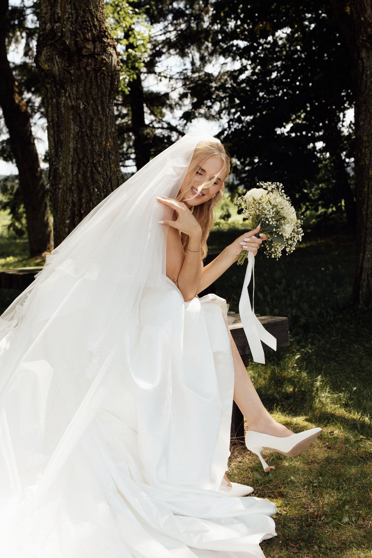 Elegant bride in white gown and veil holds floral bouquet while seated outdoors in a sunlit garden setting.