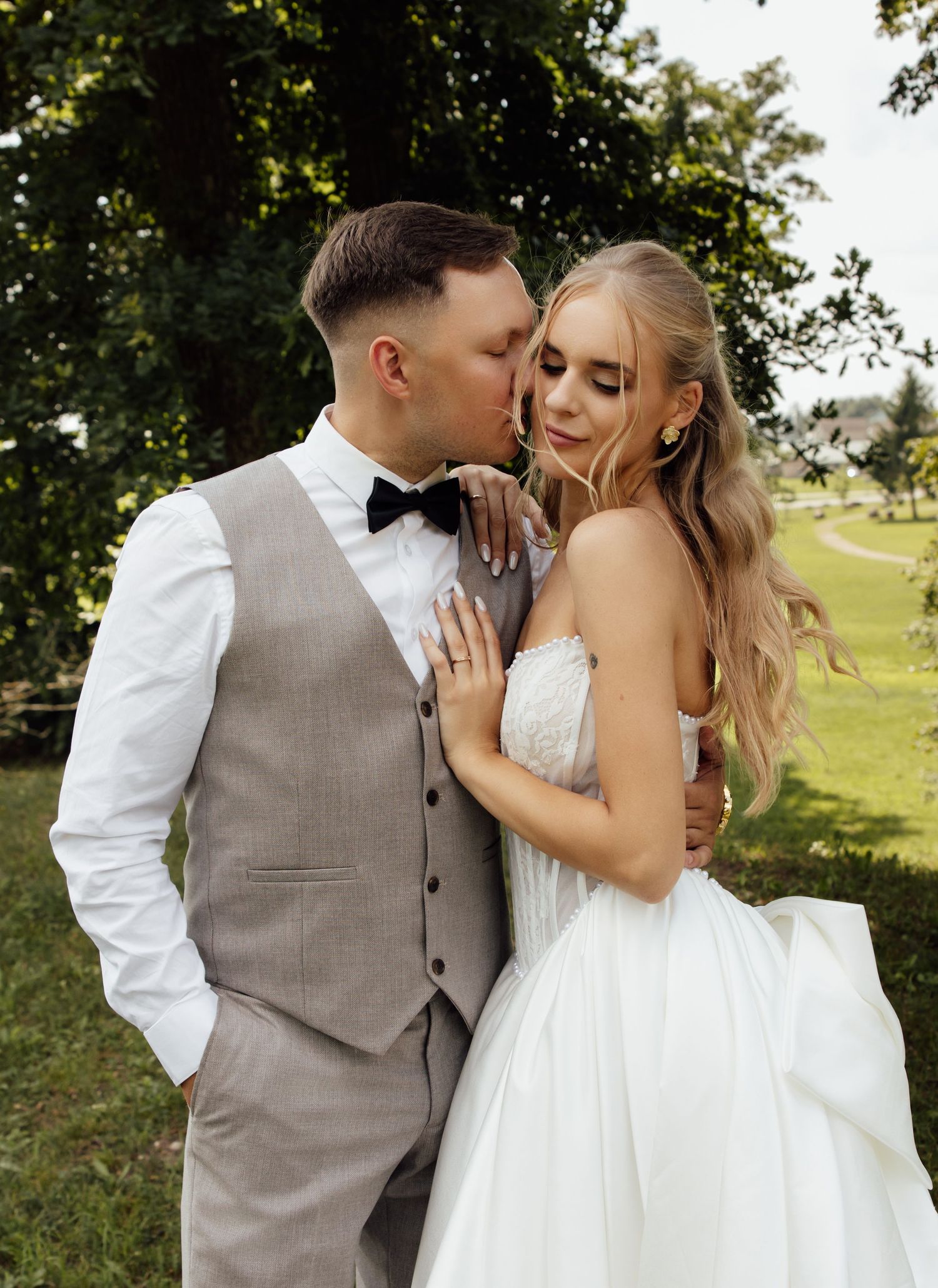 Romantic wedding couple embracing outdoors, groom in gray vest kissing bride in white gown amid lush green trees.