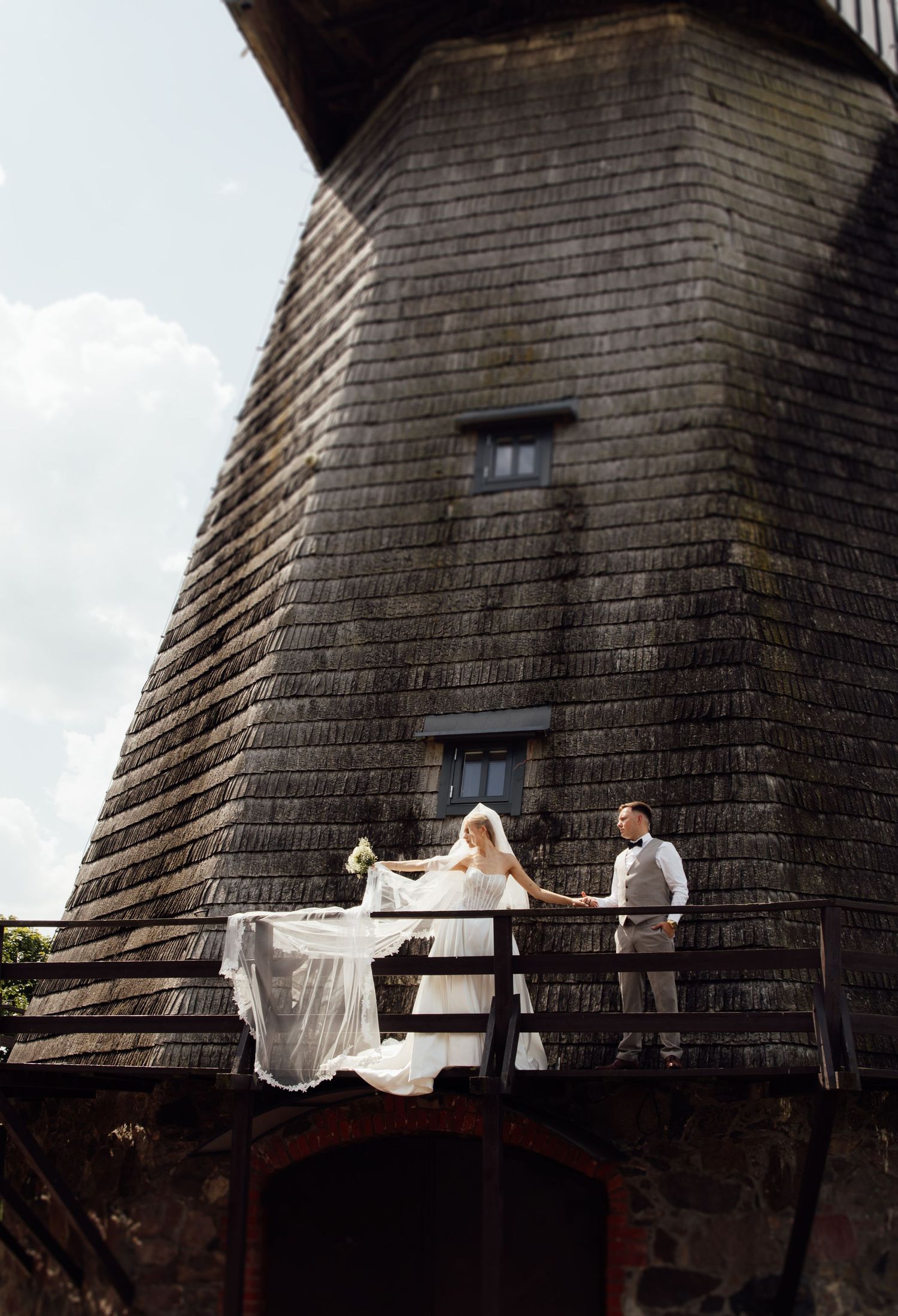Bride and groom stand on wooden balcony of historic shingled windmill tower, wedding veil flowing in breeze.