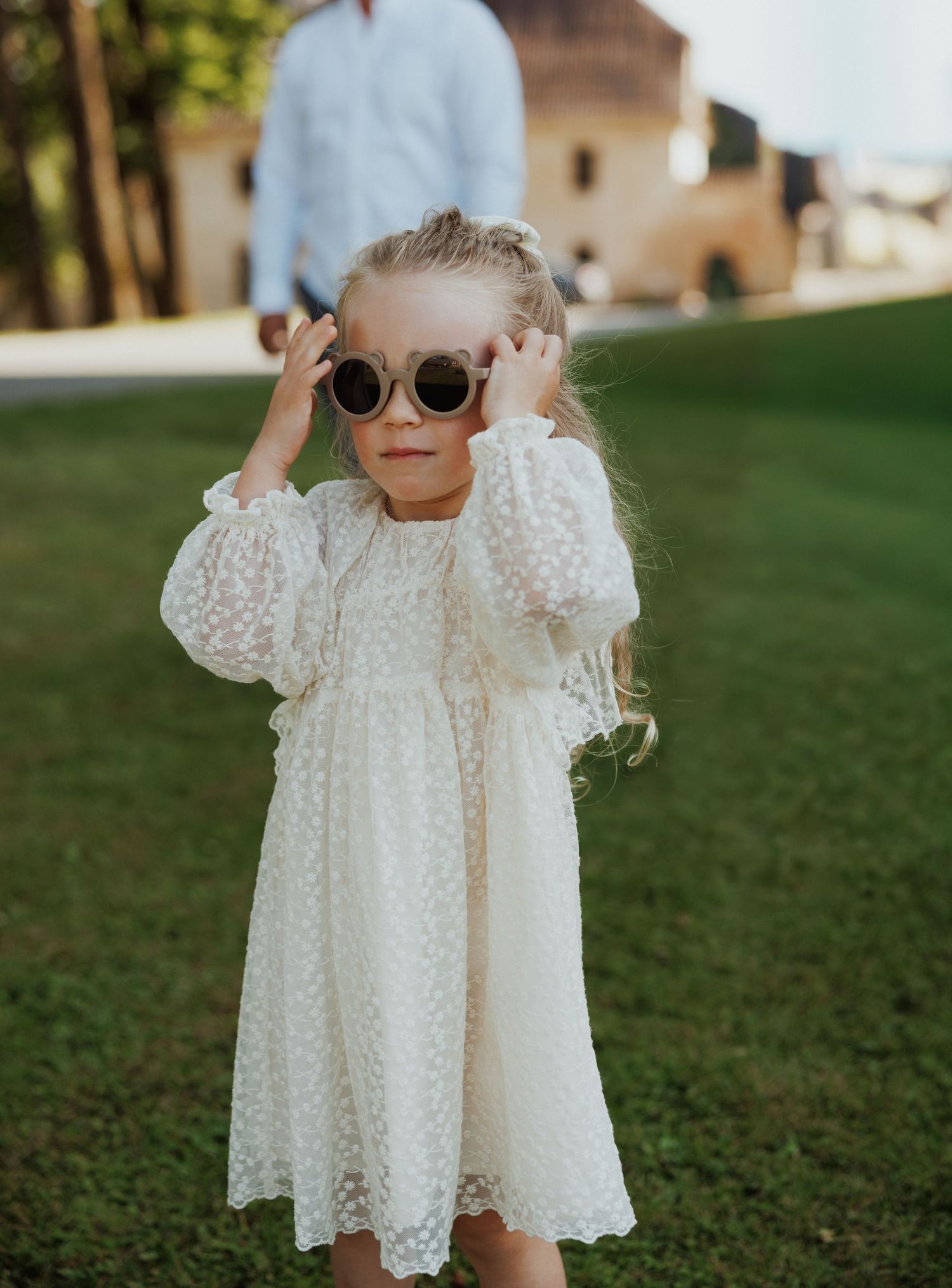 A young child in a white lace dress and round sunglasses stands on grass in warm sunlight.