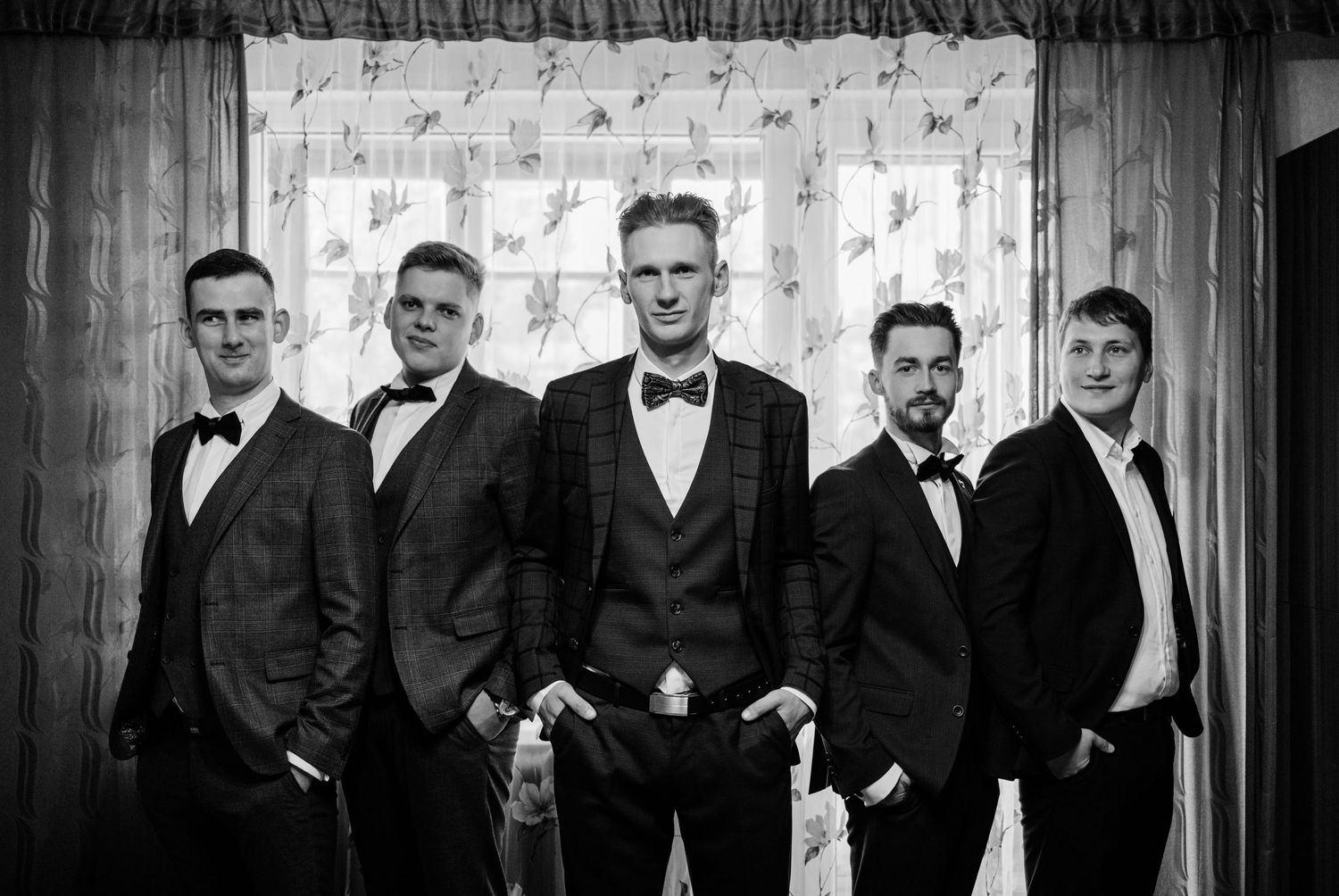 Black and white wedding party photo showing groomsmen in formal suits and bowties lined up against a dark background.