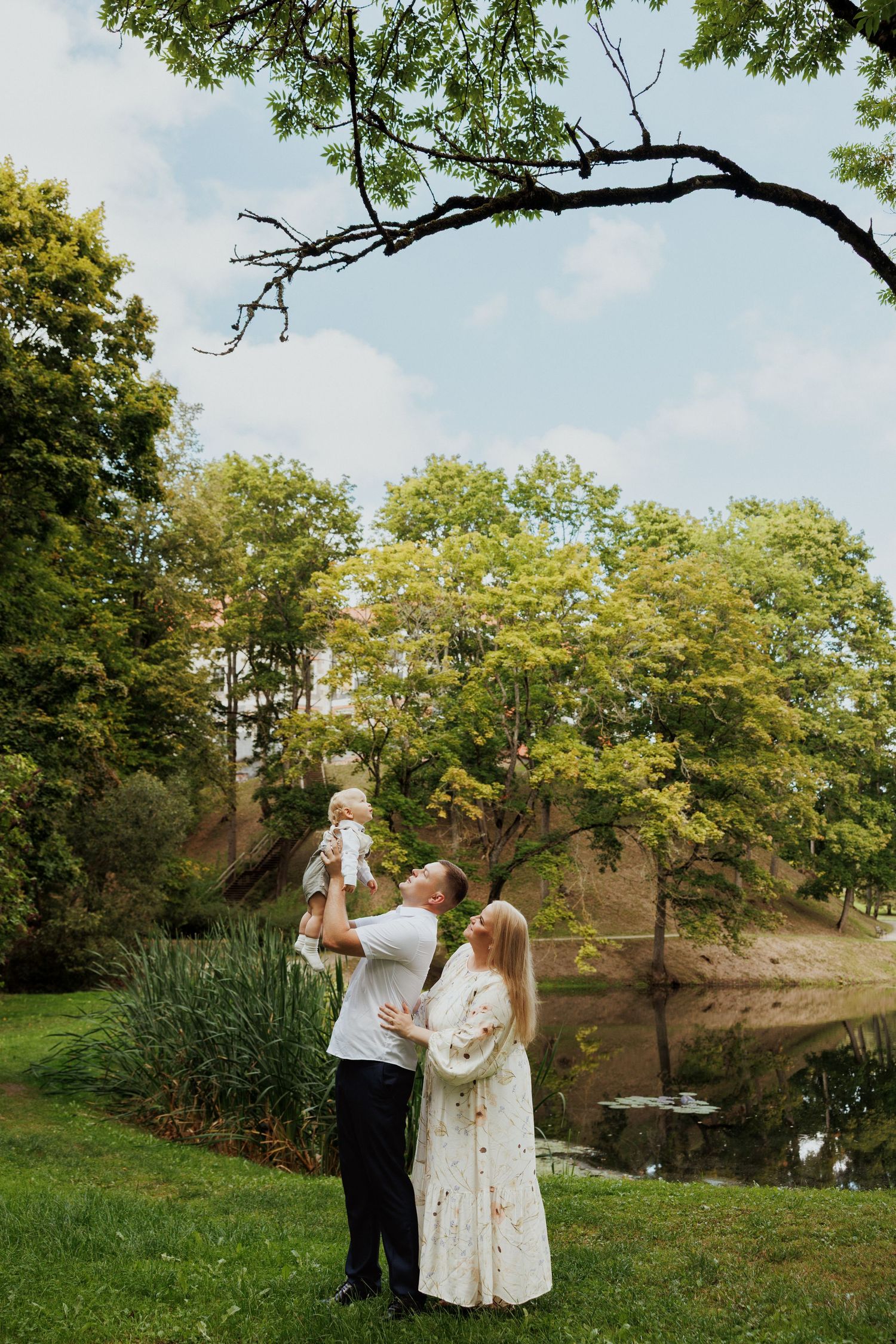 Couple shares an intimate moment together in a serene park setting with lush trees and a pond in the background.
