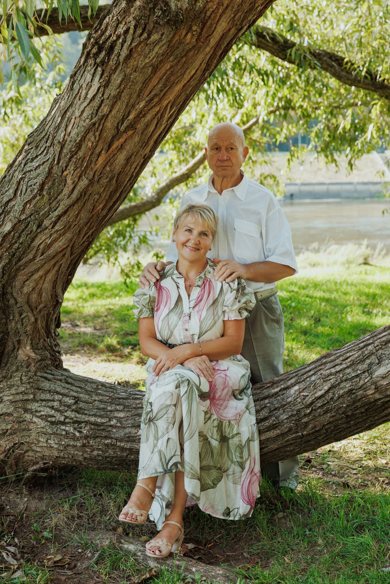 A couple poses together under a large shady tree on a sunny summer day in a park setting.