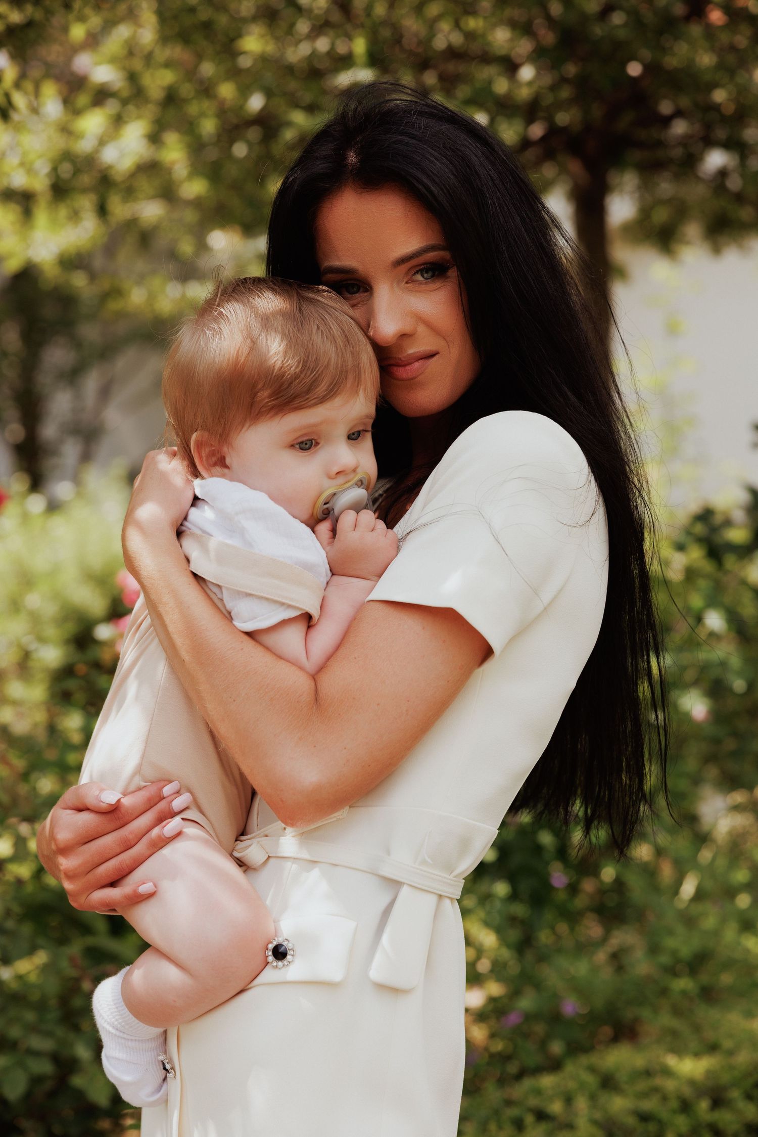 A tender moment captured outdoors with a baby in white being held against a garden background.