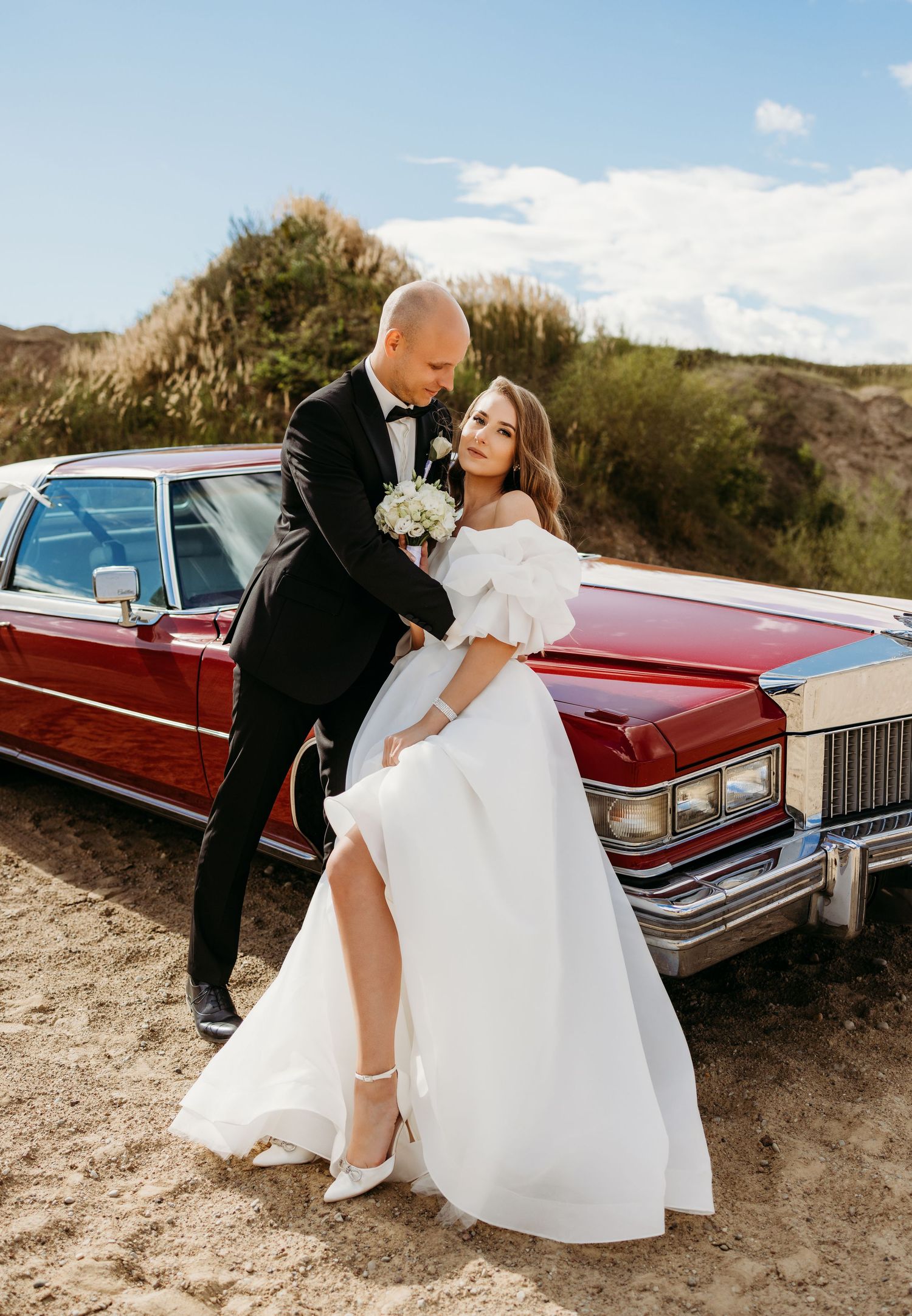 Newlyweds pose with vintage red Cadillac on desert road for romantic wedding photoshoot.