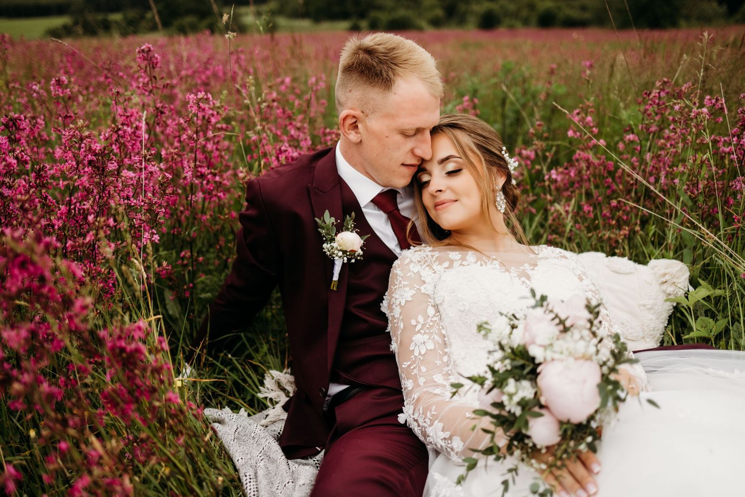 Bride in lace dress and groom in burgundy suit pose among pink flowers with white bouquet.