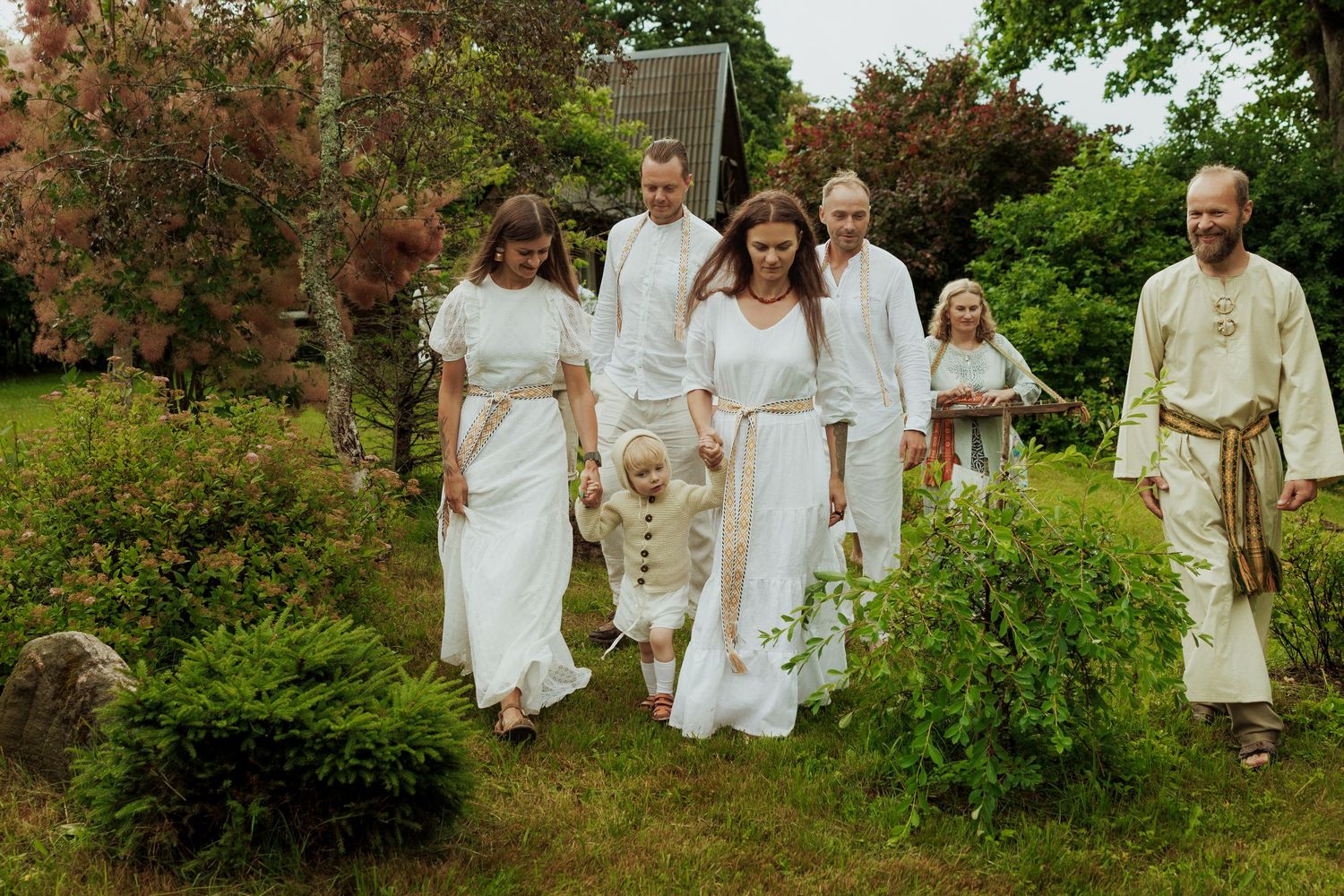 Family dressed in white traditional attire walking together through lush green garden at outdoor ceremony.
