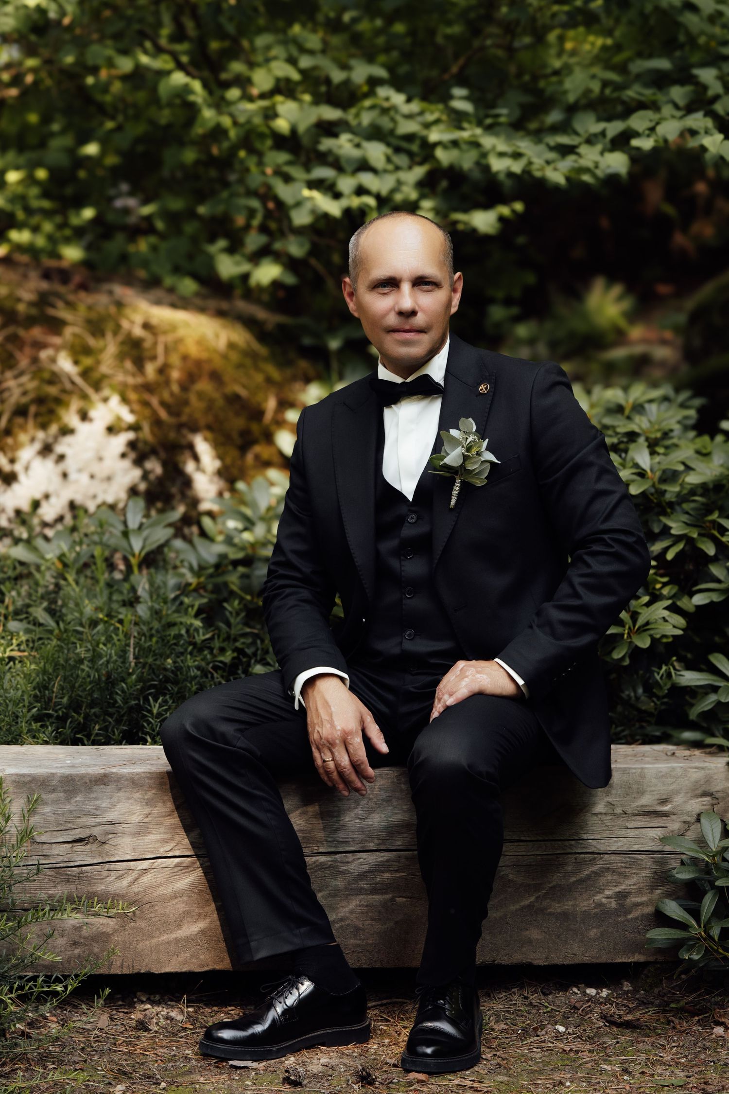 Groom in black tuxedo with boutonniere sits on wooden bench surrounded by lush green garden foliage.