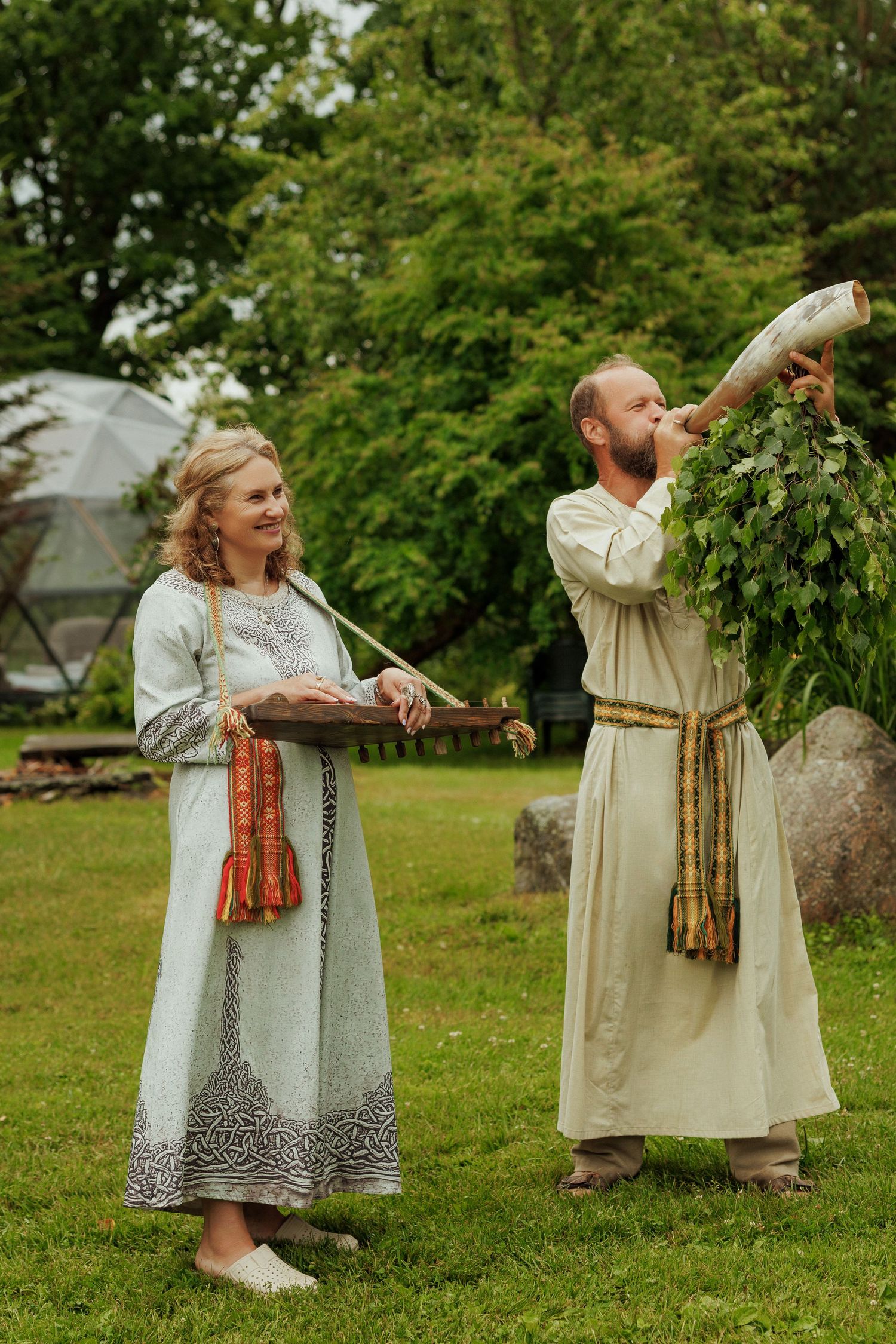 Couple in traditional folk costumes playing instruments outdoors in green garden setting.