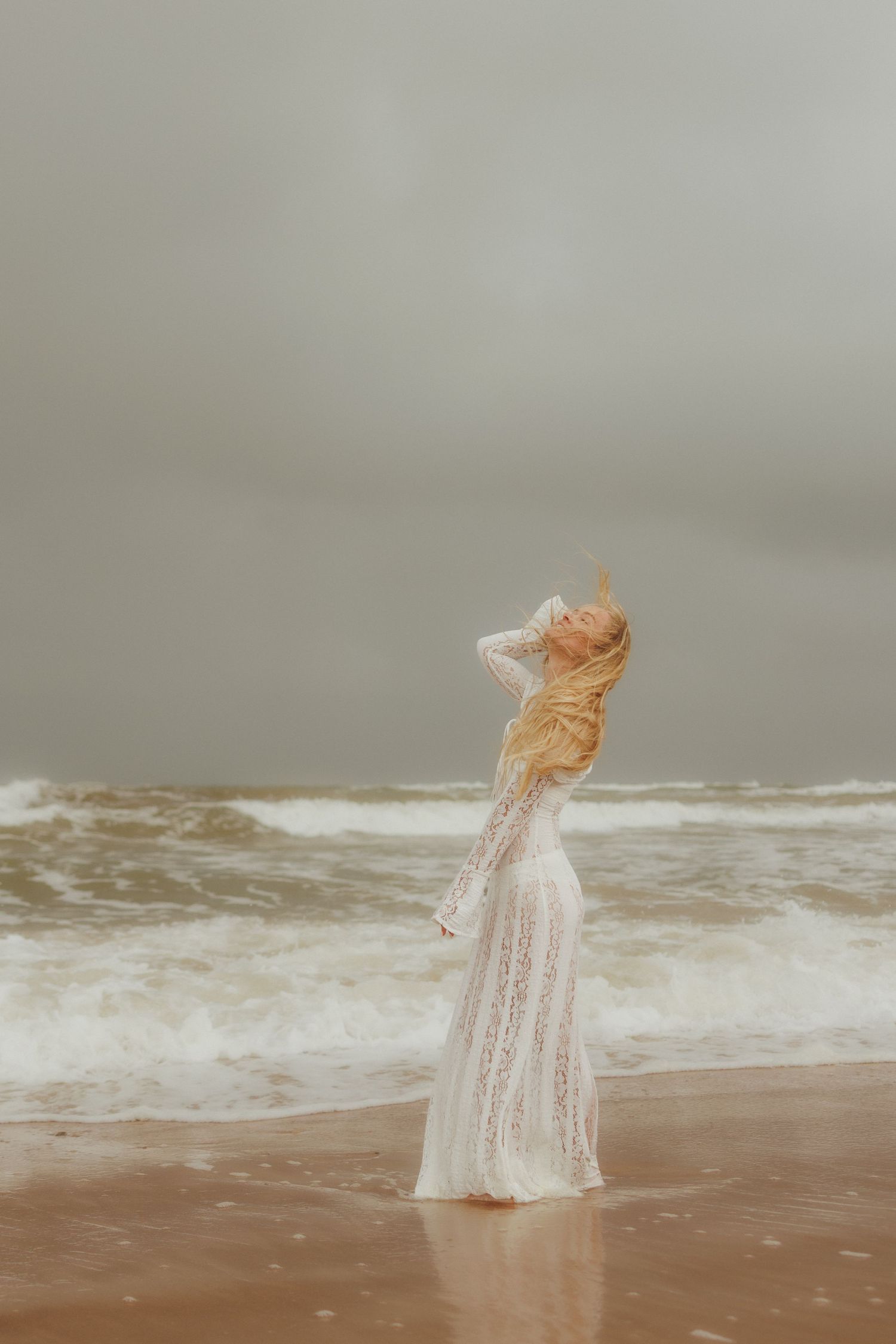 Woman in white lace gown standing on stormy beach with waves, hair blowing in wind on overcast day.