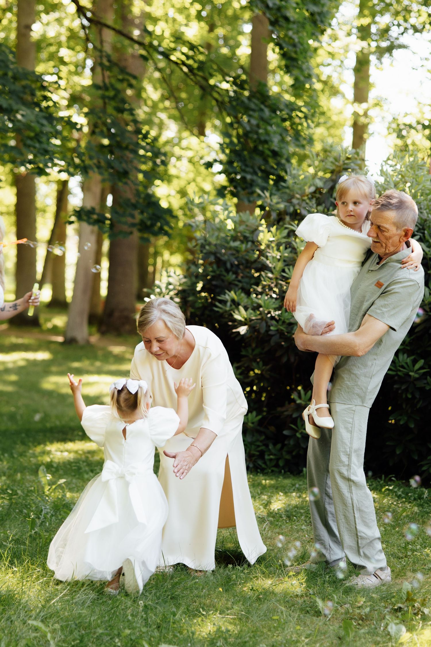 Grandparents playing with two young girls in white dresses outdoors in a sunny green park.