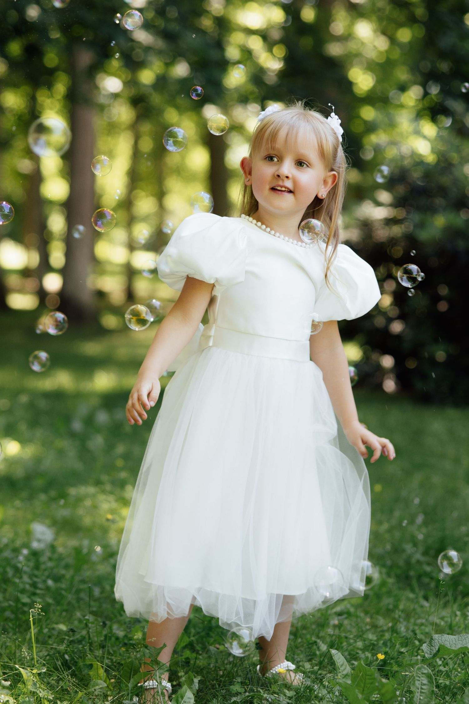 Young girl in white tulle dress with puff sleeves playing among soap bubbles in a sunny green park.