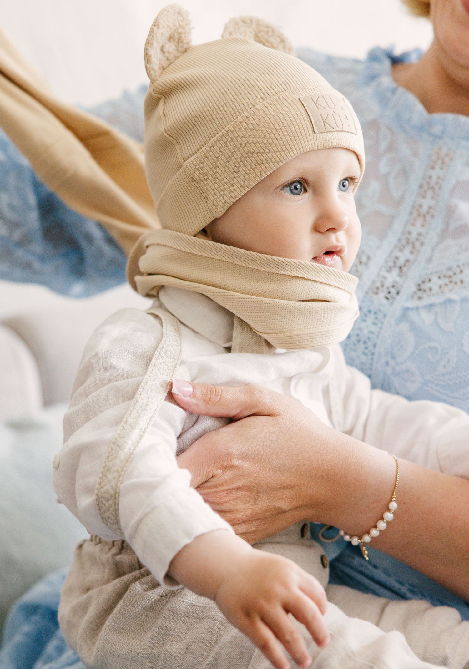 A small child wearing a beige knit hat with ears and matching scarf in a cozy indoor setting.