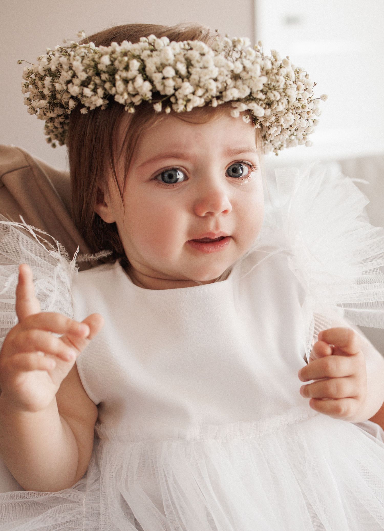 Baby wearing a white dress and delicate baby's breath flower crown looking up with raised finger.