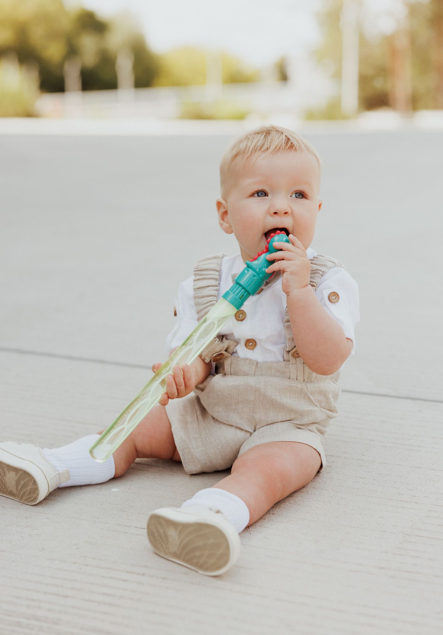 Baby in beige romper lying on white surface playing with a teal colored toy.