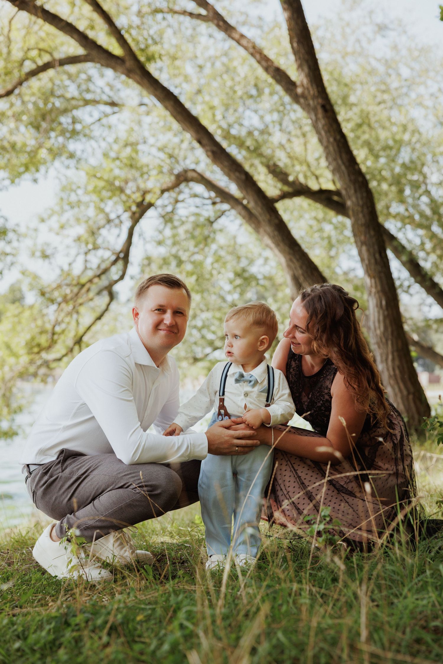 Family enjoying a tender moment together while sitting on grassy hill under trees on a sunny day.