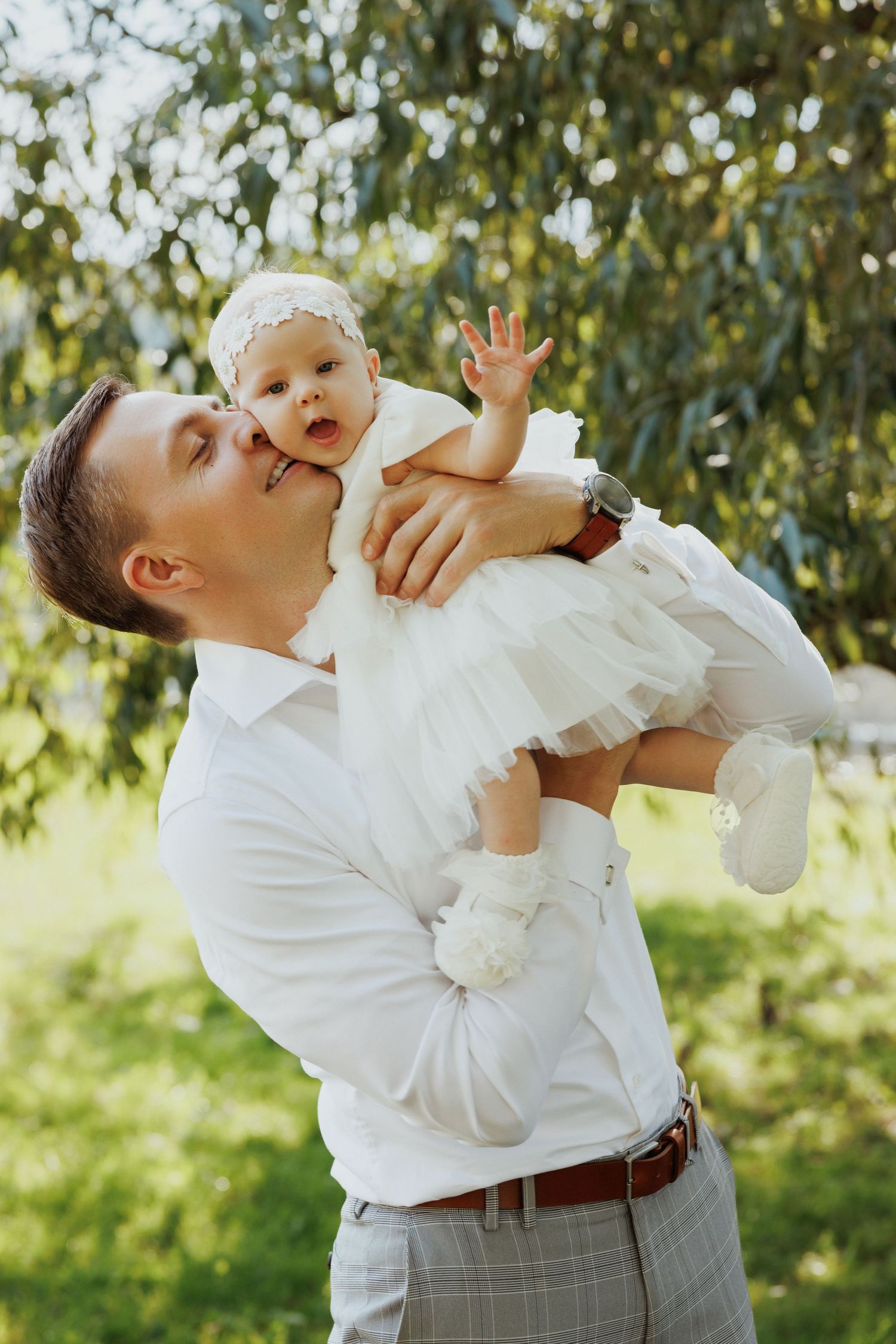 Three heartwarming moments of a parent lifting and kissing a baby in white dress outdoors against green foliage.